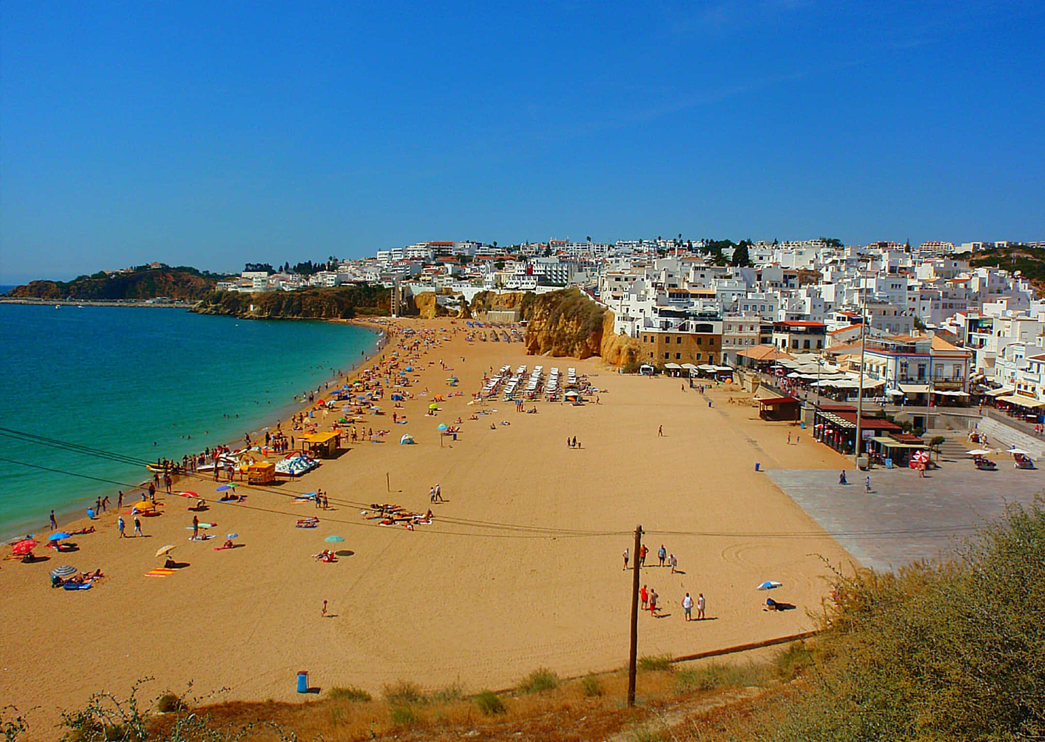 Portuguese Beach 桌布 壁紙 — 一個充滿活力的陽光海灘，在清澈的藍天下擠滿遊客，白色懸崖建築俯瞰著碧綠的海水。