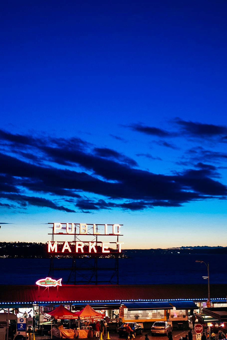 Pike Place Market 桌布 壁紙 — 一個充滿活力的霓虹燈「PUBLIC MARKET」標誌，在深藍色的暮色天空和散落的雲朵下，於繁忙的海濱攤位區域上方閃耀。