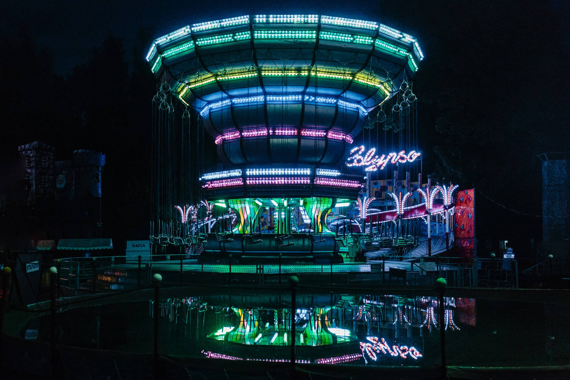 Night Aesthetic 桌布 壁紙 — 一個充滿活力的霓虹燈旋轉木馬在夜晚閃耀，色彩繽紛的燈光倒映在下方平靜的水池中。