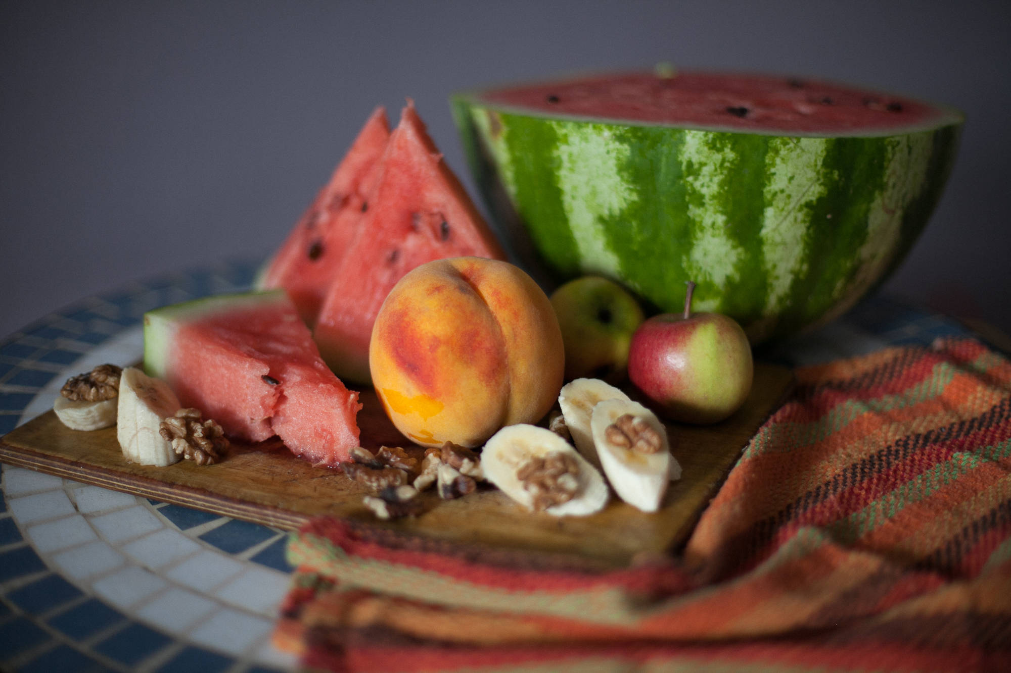 Fruits 桌布 壁紙 — 一個充滿活力的靜物擺設，將切片西瓜、桃子、香蕉和蘋果放在鋪有圖案布料的木板上。