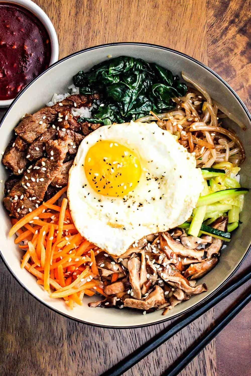 烹飪 桌布 壁紙 — 一個充滿活力的韓國石鍋拌飯碗，上面放著太陽蛋、芝麻和色彩繽紛的蔬菜，放在木桌上。