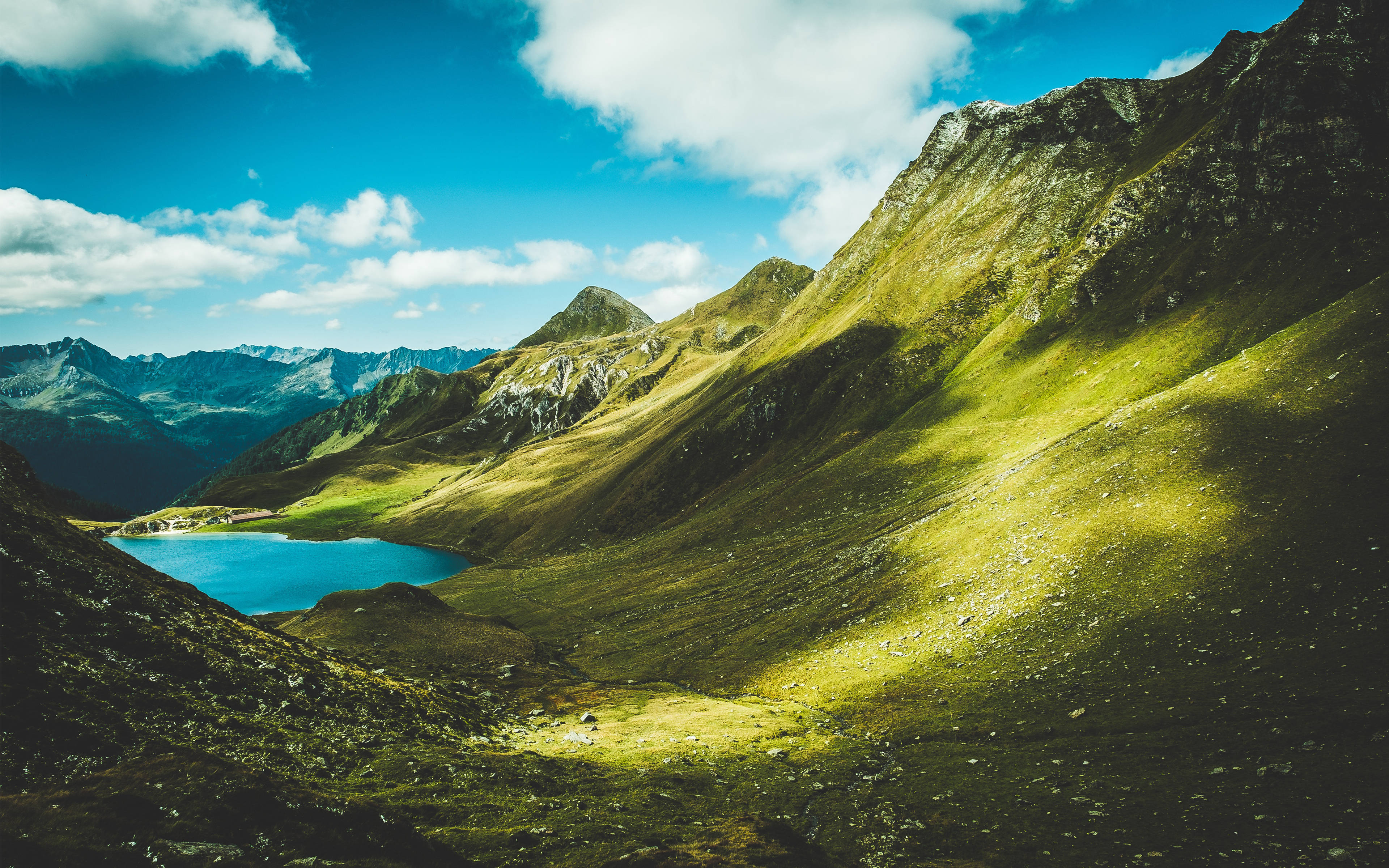 4K Summer 桌布 壁紙 — 一個充滿活力的高山湖泊，坐落在陽光明媚的綠色山脈中，上方是明亮的藍天和蓬鬆的白雲。