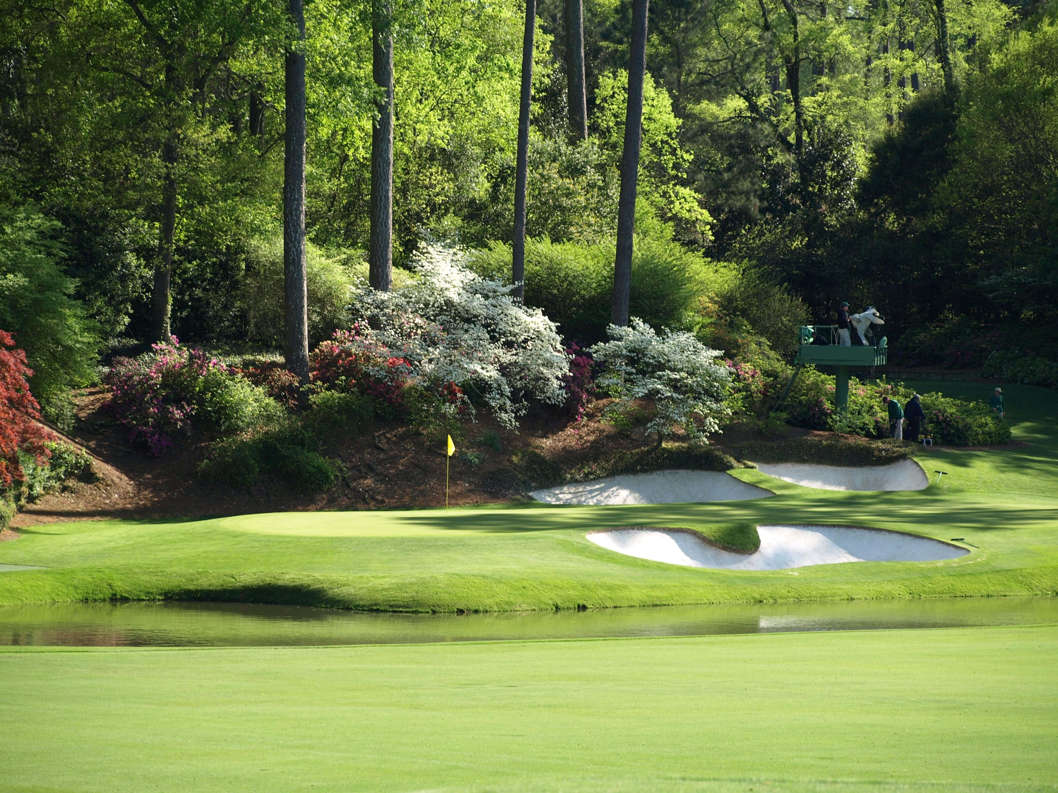 Augusta National iPhone 桌布 壁紙 — 一個充滿活力的高爾夫果嶺，周圍環繞著沙坑和色彩繽紛的花卉灌木，在明亮的陽光下。