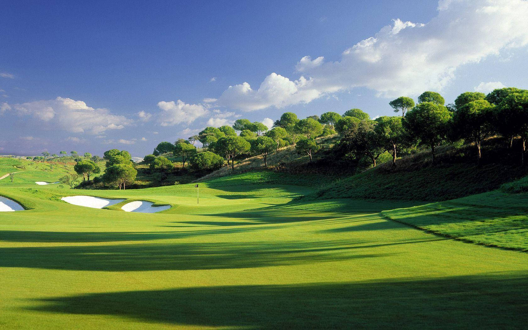 Golfing 桌布 壁紙 — 一個充滿活力的高爾夫球場，擁有鬱鬱蔥蔥的綠色球道、沙坑和樹木繁茂的山丘，在明亮的藍天和白雲下。