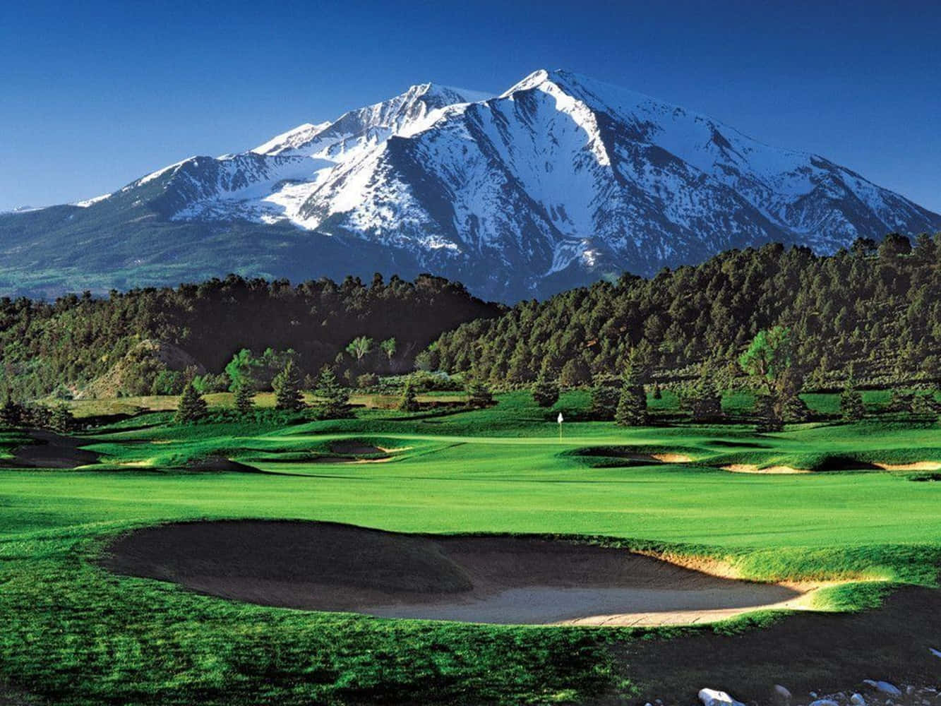 Awesome Golf 桌布 壁紙 — 一個充滿活力的高爾夫球場，擁有鬱鬱蔥蔥的綠色球道和沙坑，在晴朗的藍天下，在雄偉的雪山前延伸。