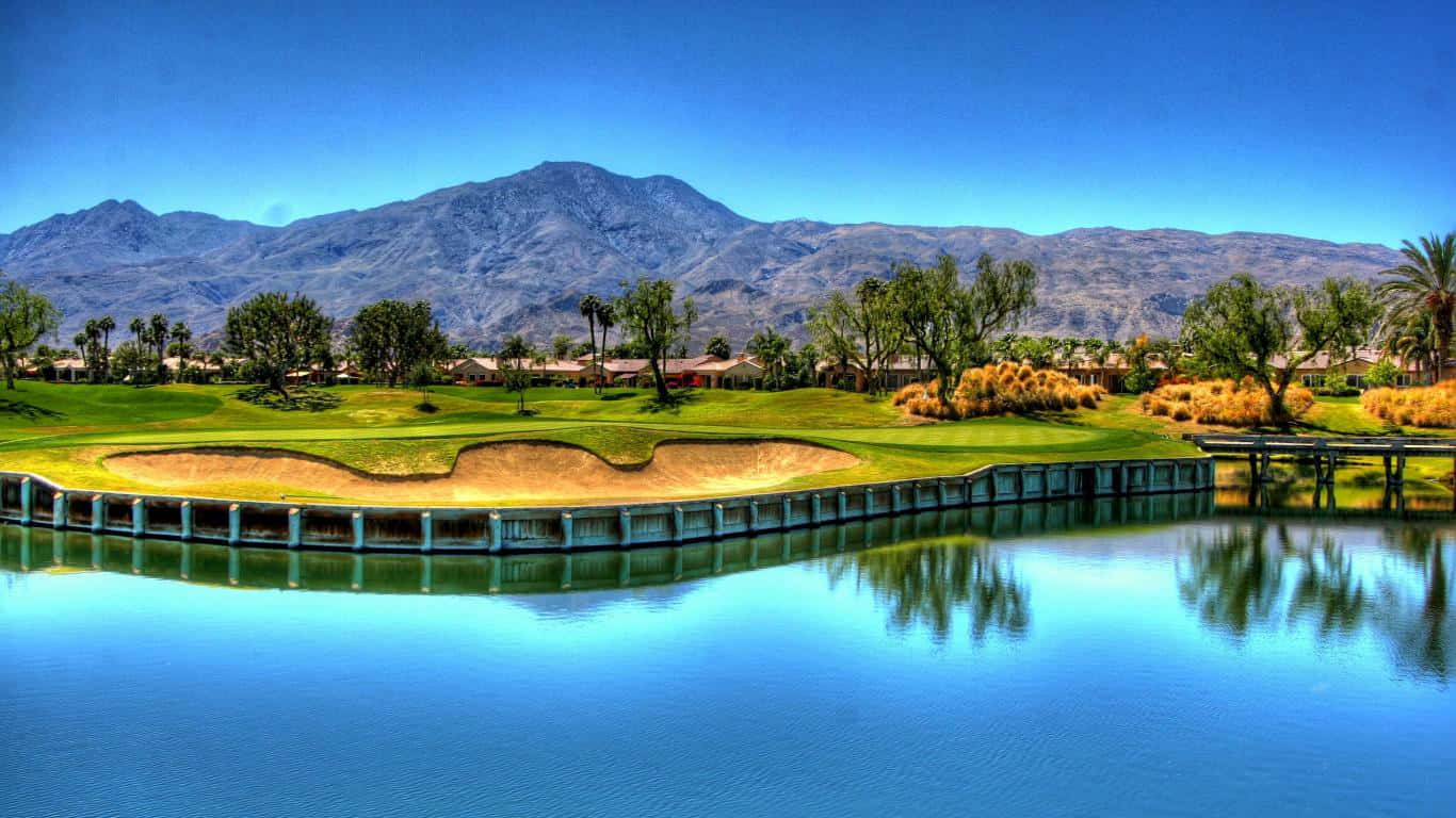 1366x768 Golf Course 桌布 壁紙 — 一個充滿活力的高爾夫球場，設有大型水障礙和沙坑，背景是晴朗的藍天和山脈。