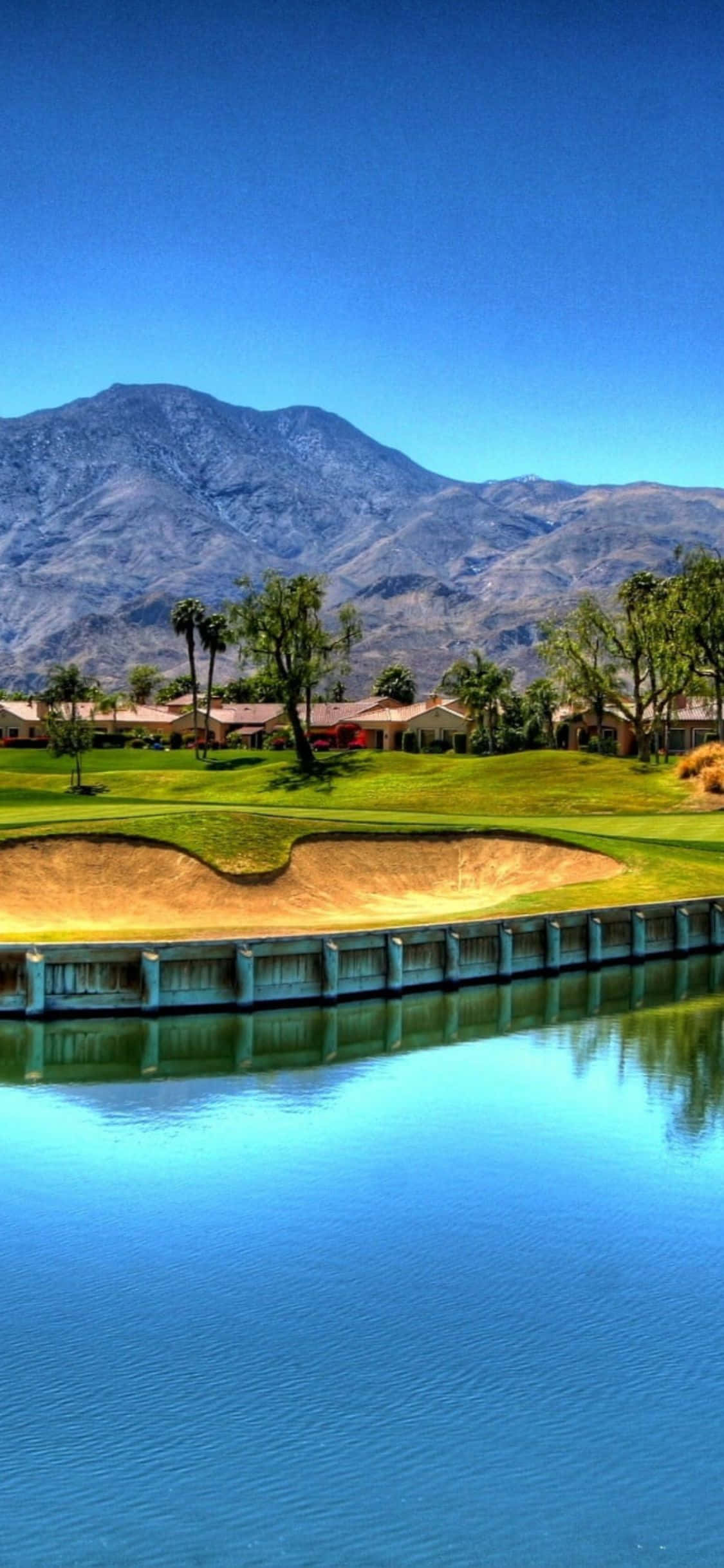 iPhone X Golf Course 桌布 壁紙 — 一個充滿活力的高爾夫球場，設有水障礙和沙坑，在晴朗的藍天下延伸至崎嶇的山脈前。