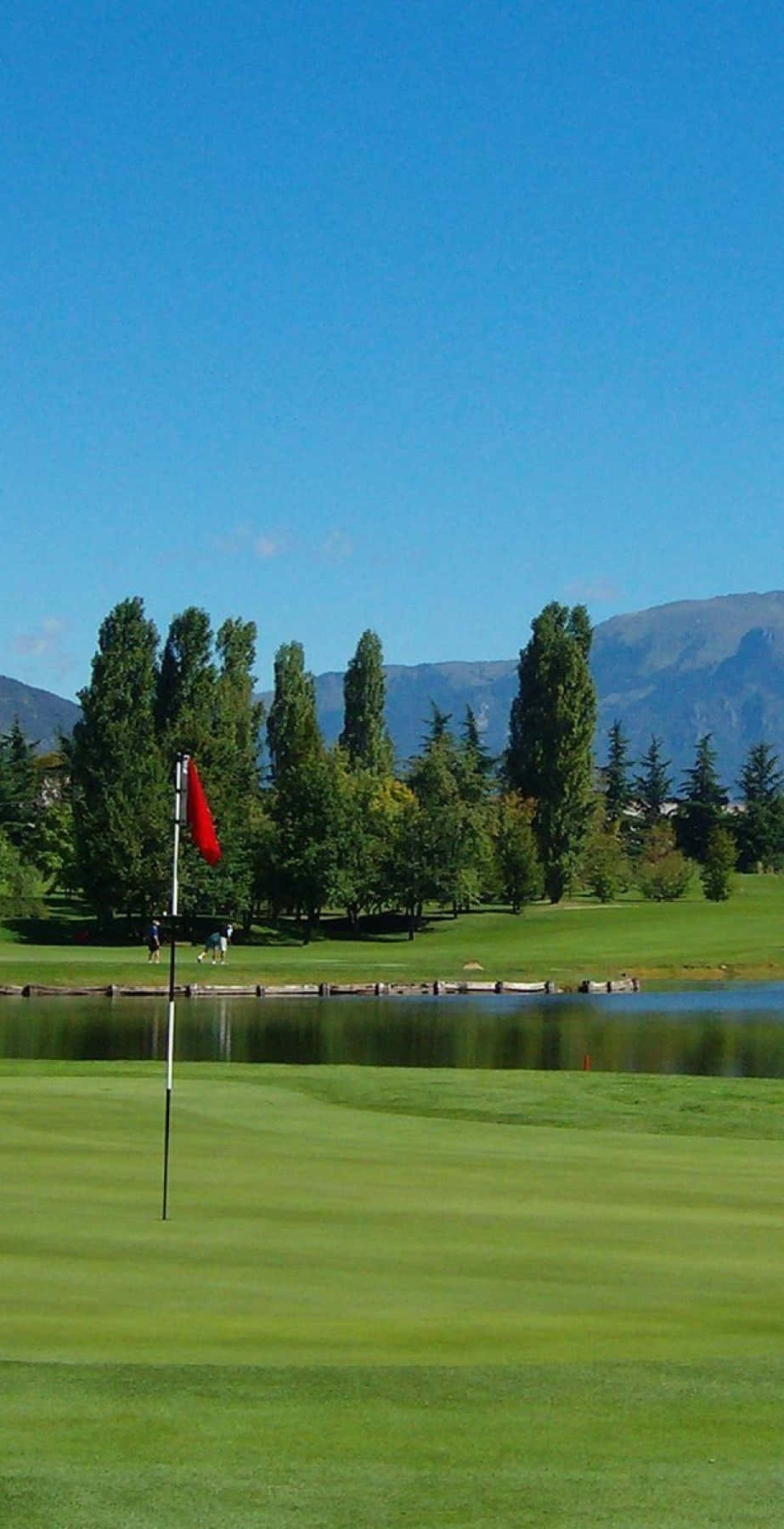 Pixel 3 Golf Course 桌布 壁紙 — 一個充滿活力的高爾夫球場果嶺，靠近平靜的池塘，周圍有高大的樹木和遠處的山脈，在晴朗的藍天下。