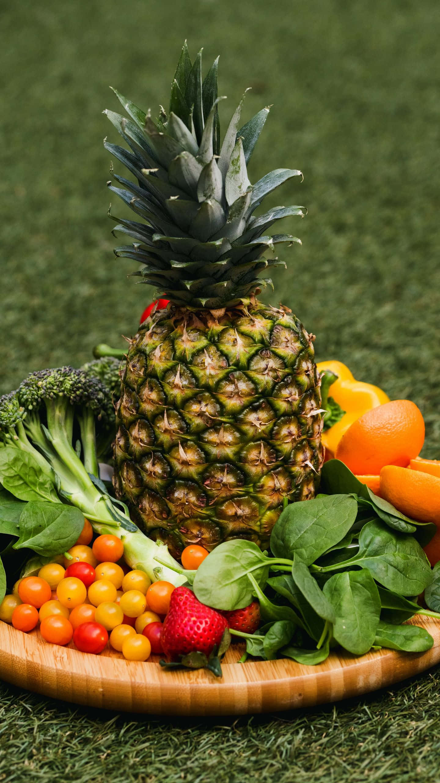 Pineapple 桌布 壁紙 — 一個充滿活力的鳳梨，被櫻桃番茄、草莓、菠菜和柳橙圍繞，放在綠色草地上的木盤上。
