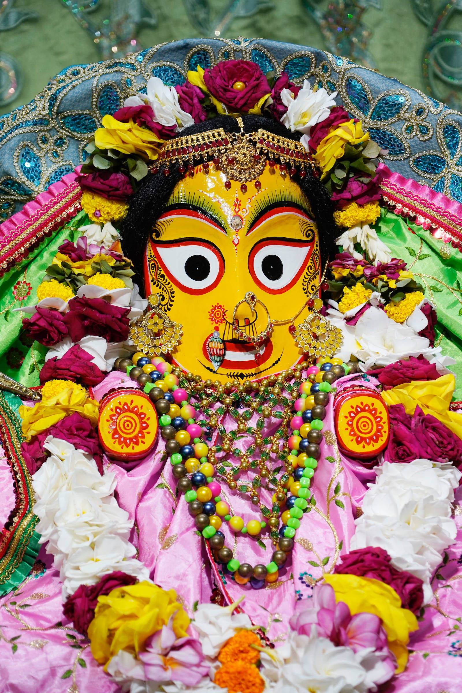 Shankar Bhagwan 桌布 壁紙 — 一個充滿活力的黃臉印度教女神像，裝飾著色彩繽紛的花朵、金飾和粉紅色布料，在節日環境中。