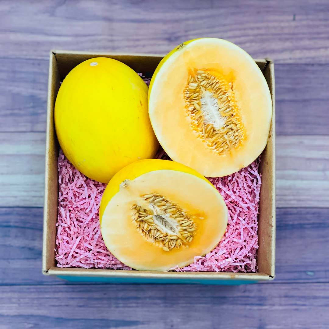 Fruit 桌布 壁紙 — 一個充滿活力的黃色哈密瓜切半，露出多汁的橙色果肉和種子，放在粉紅色碎紙中，置於紙箱內。