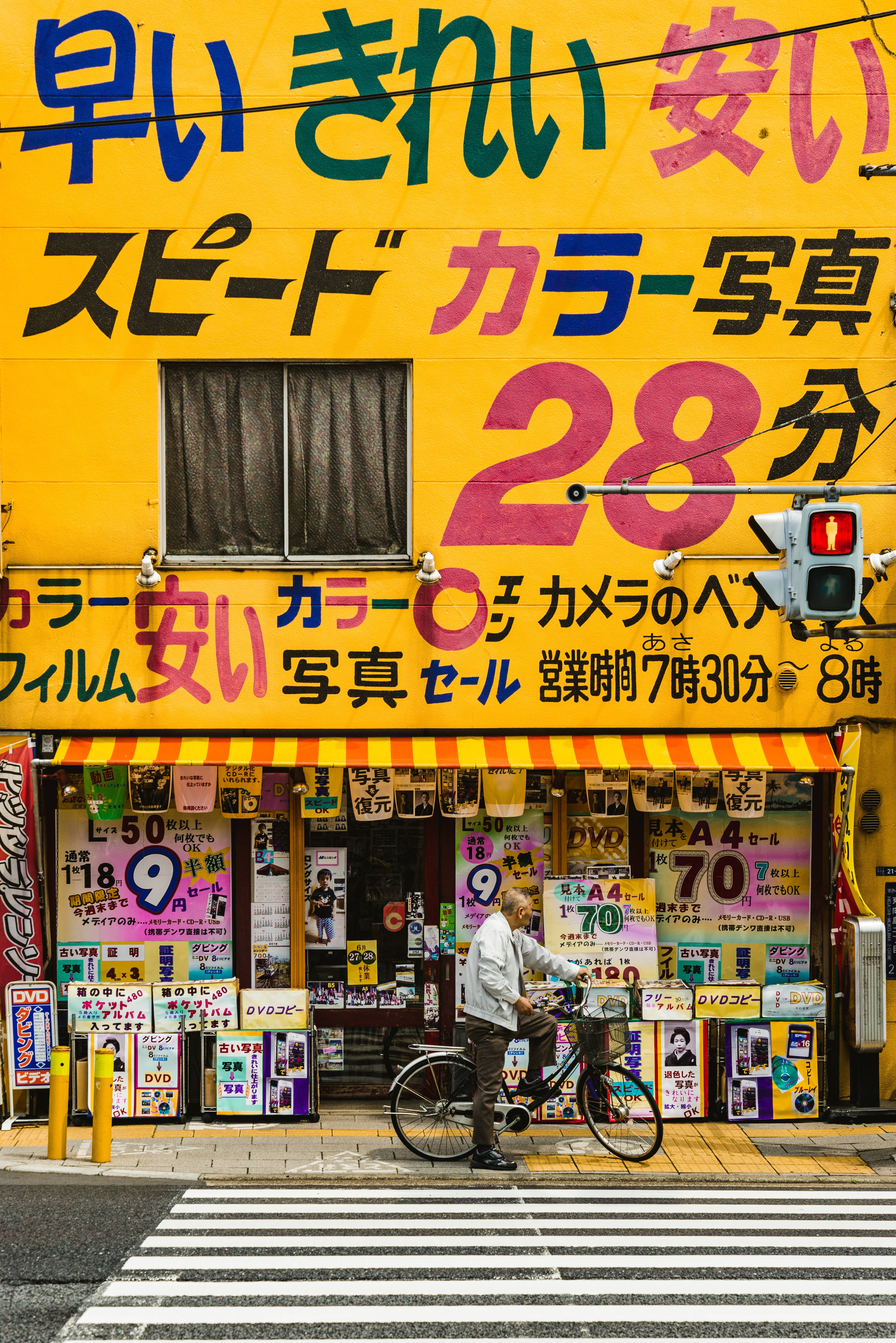 動漫角色 桌布 壁紙 — 一個充滿活力的黃色日本照相館招牌，帶有粗體彩色文字，以及一個騎腳踏車的男人在城市街道上經過。