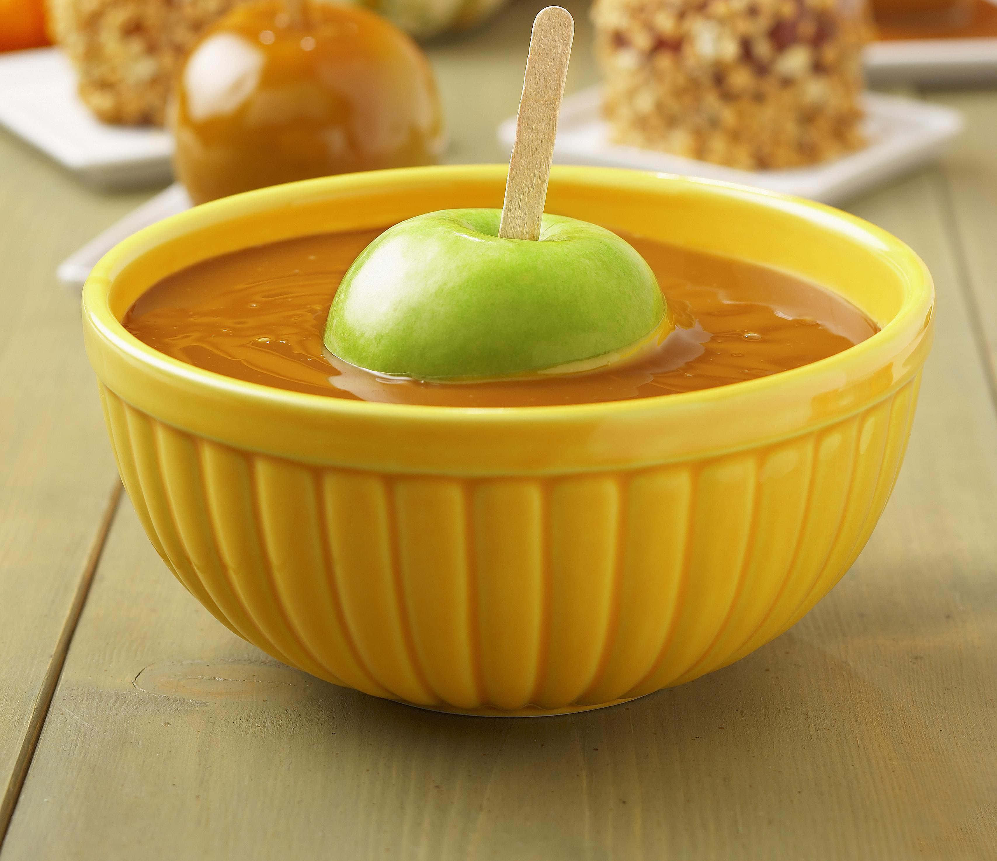 Caramel Apples 桌布 壁紙 — 一個充滿活力的黃色條紋碗盛載著閃亮的焦糖醬，上面插著一支沾滿焦糖醬的青蘋果棒，放在木桌上。