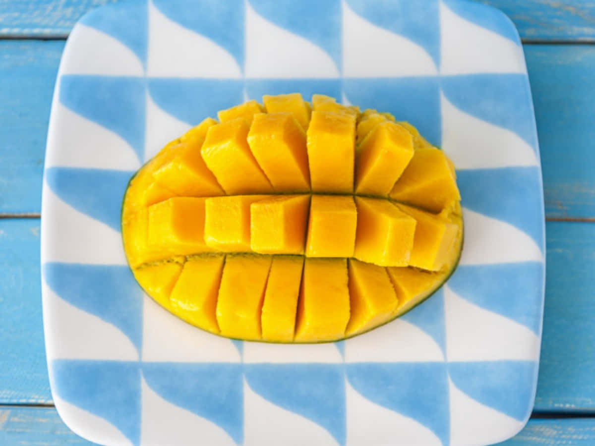 Mango 桌布 壁紙 — 一個充滿活力的黃色芒果切成整齊的方塊，放在一個藍白格子盤上，置於質樸的藍色木質表面上。