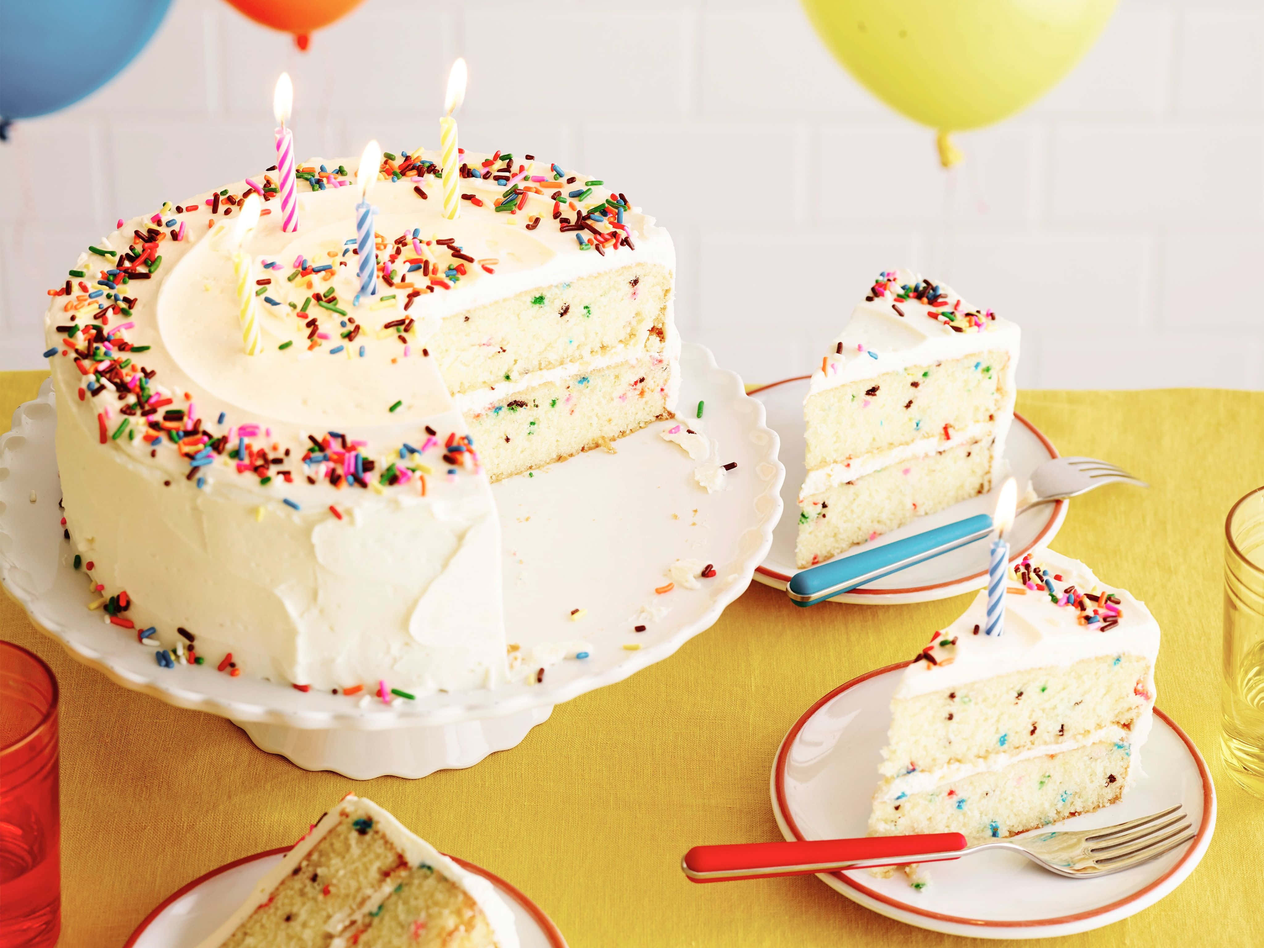 Birthday 桌布 壁紙 — 一個充滿節慶氣氛的funfetti生日蛋糕，上面有彩色糖屑和點燃的蠟燭，放在黃色桌布上，還有氣球和切好的蛋糕片。