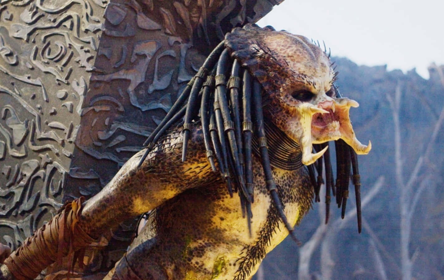 Predator 桌布 壁紙 — 一個兇猛的終極戰士戰士張開鋸齒狀的牙齒咆哮，背景是雕刻的石頭和光禿的樹木。