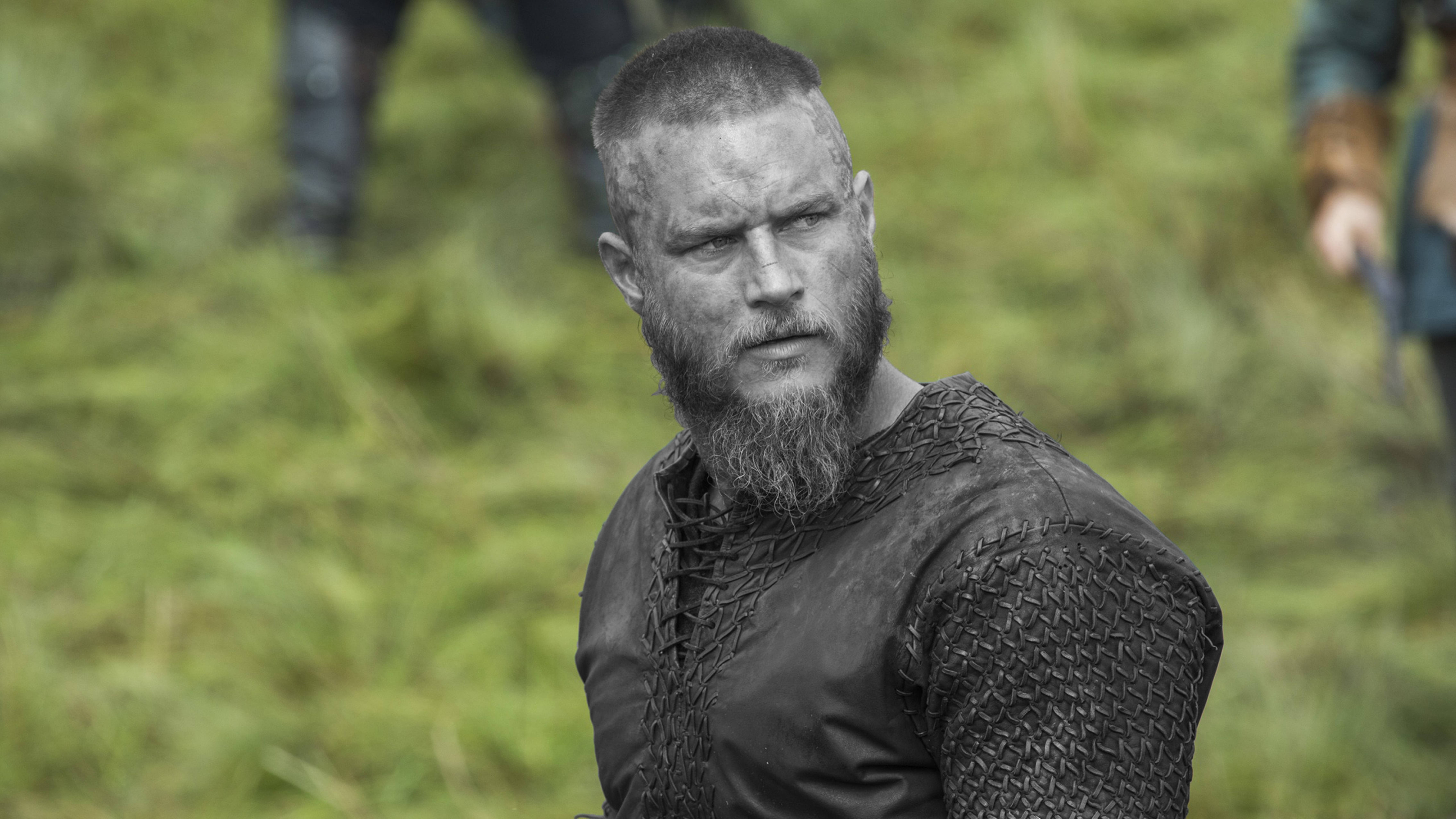 Vikings 桌布 壁紙 — 一個兇猛的鬍鬚維京戰士身穿鎖子甲，專注地凝視著模糊的綠色草地。