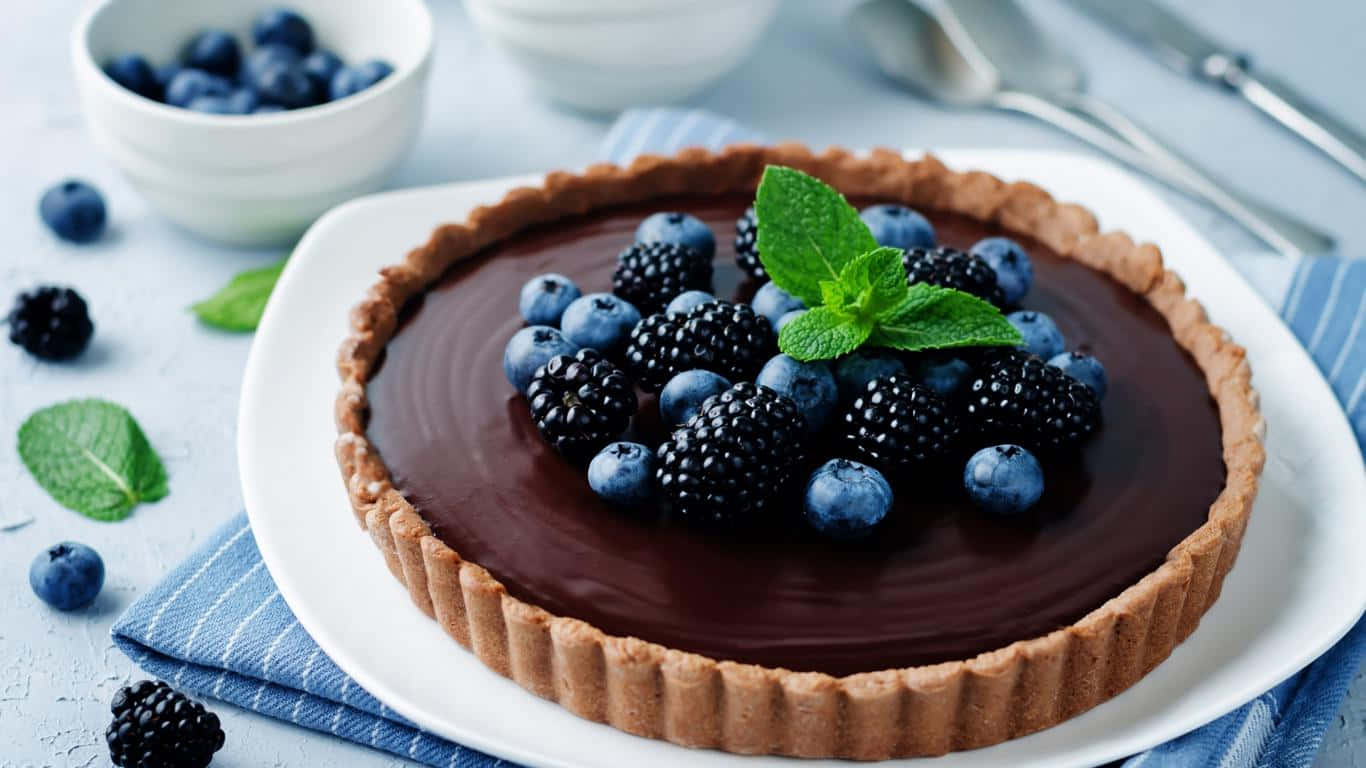 1366x768 Desserts 桌布 壁紙 — 一個光滑的巧克力塔，上面鋪滿新鮮藍莓、黑莓和薄荷葉，放在一個配有藍色條紋餐巾的白色盤子上。
