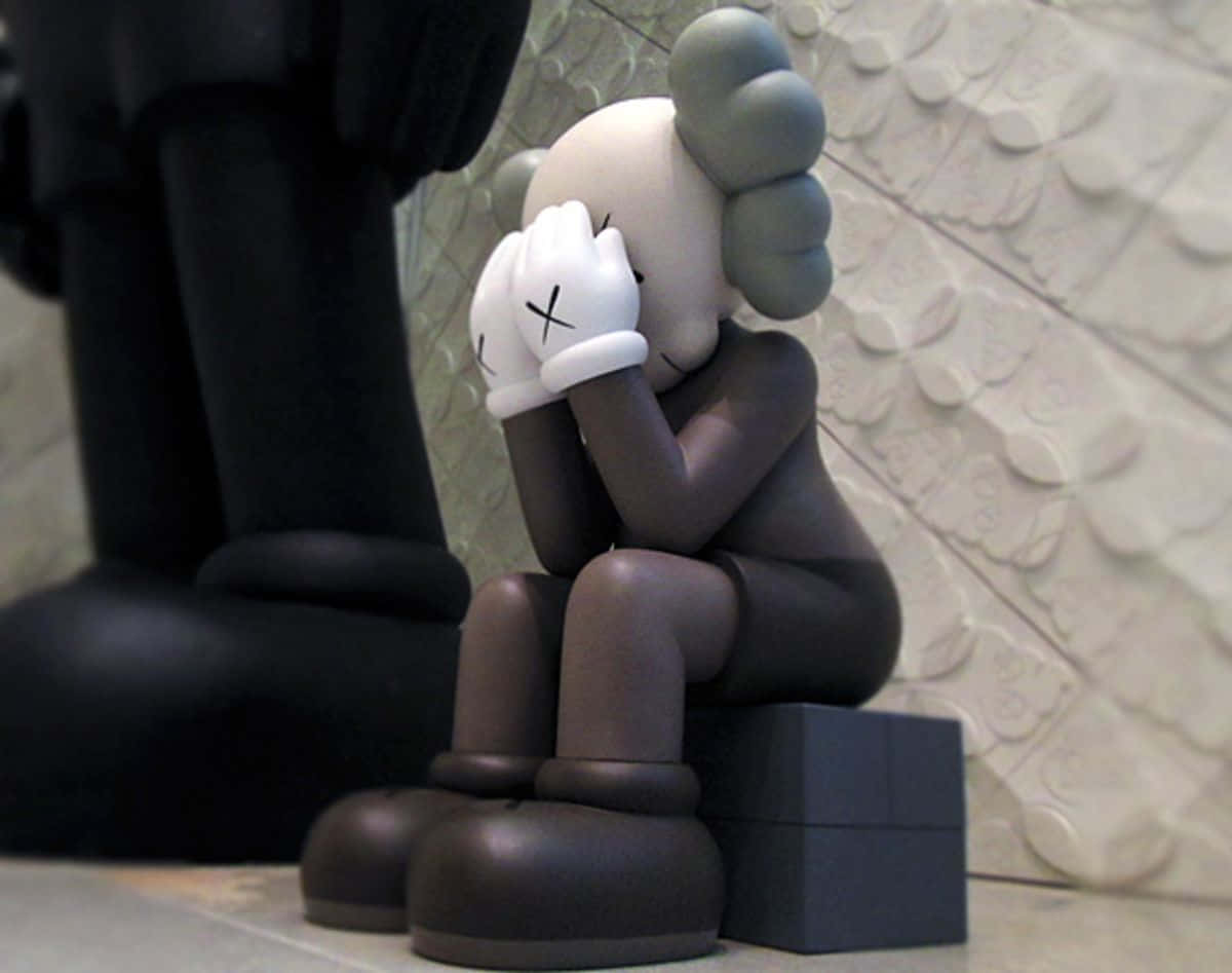 Kaws Astro Boy 桌布 壁紙 — 一個光滑的棕色KAWS雕塑，雙手掩面，蜷縮著坐在有紋理的白色牆壁前。