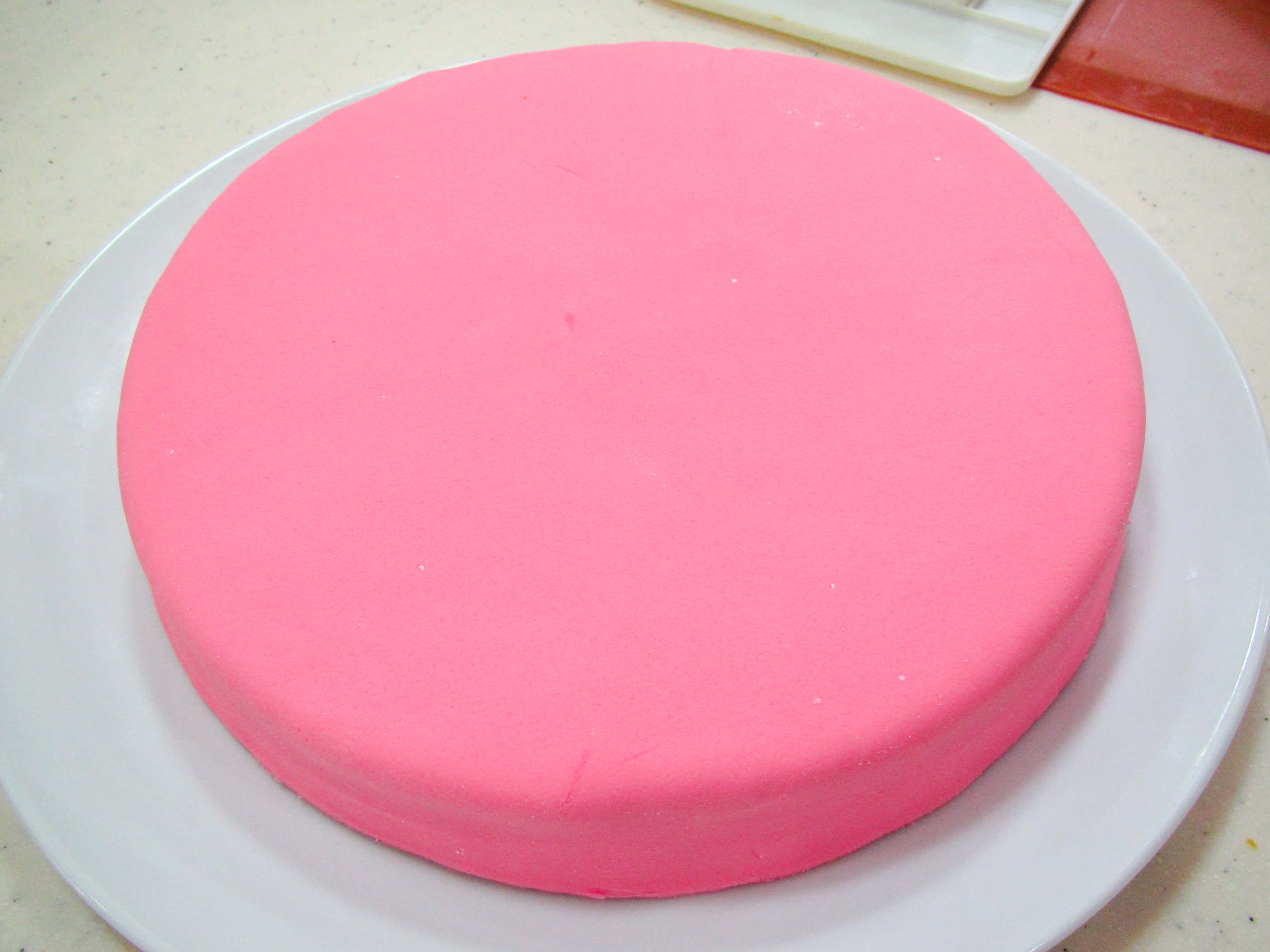 Cake Fondant 桌布 壁紙 — 一個光滑的淺粉紅色糖霜蛋糕放在白色盤子中央，置於淺色斑點檯面上。