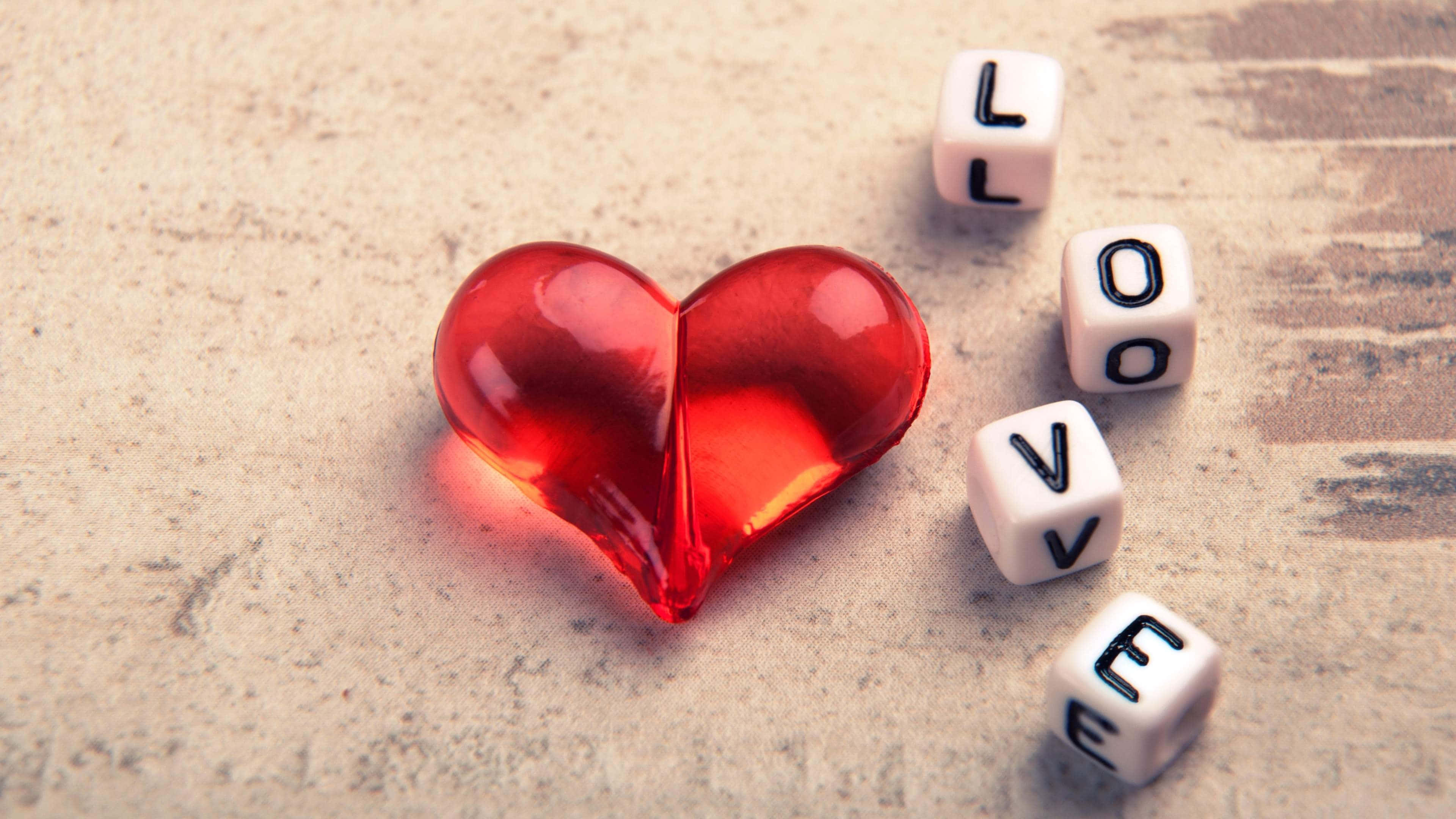 4K Love 桌布 壁紙 — 一個光滑的紅色心形物體放在白色骰子旁邊，骰子在紋理米色表面上拼出 love。