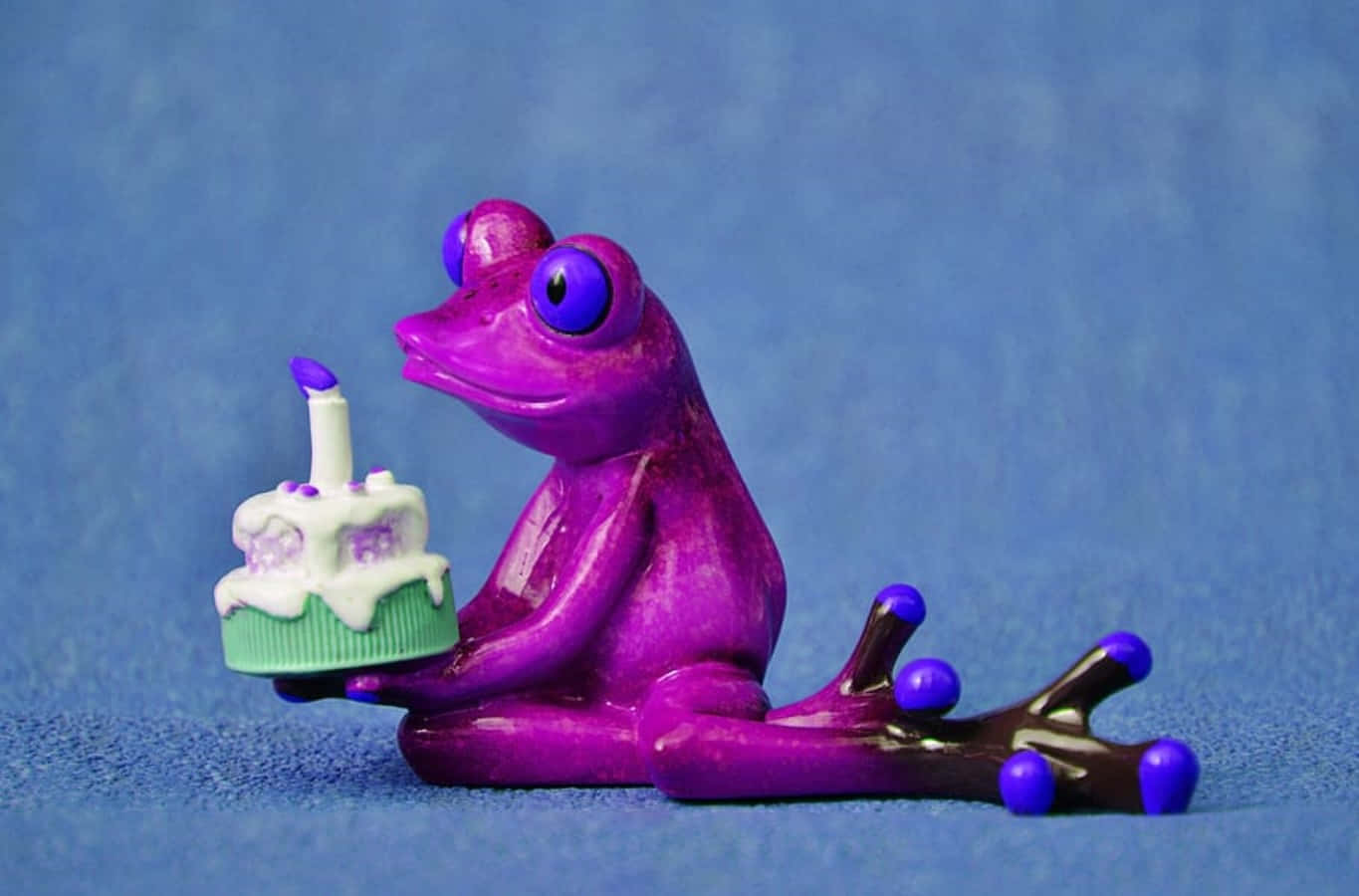 Birthday Cake 桌布 壁紙 — 一個光滑的紫色青蛙公仔坐著，手持一個插著一支點燃蠟燭的小蛋糕，背景是帶紋理的藍色。