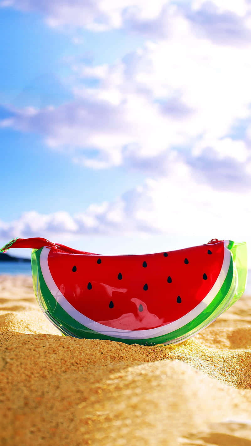 Watermelon iPhone 桌布 壁紙 — 一個光滑的西瓜形狀小袋放在金色沙灘上，上方是晴朗藍天和 fluffy 雲朵。