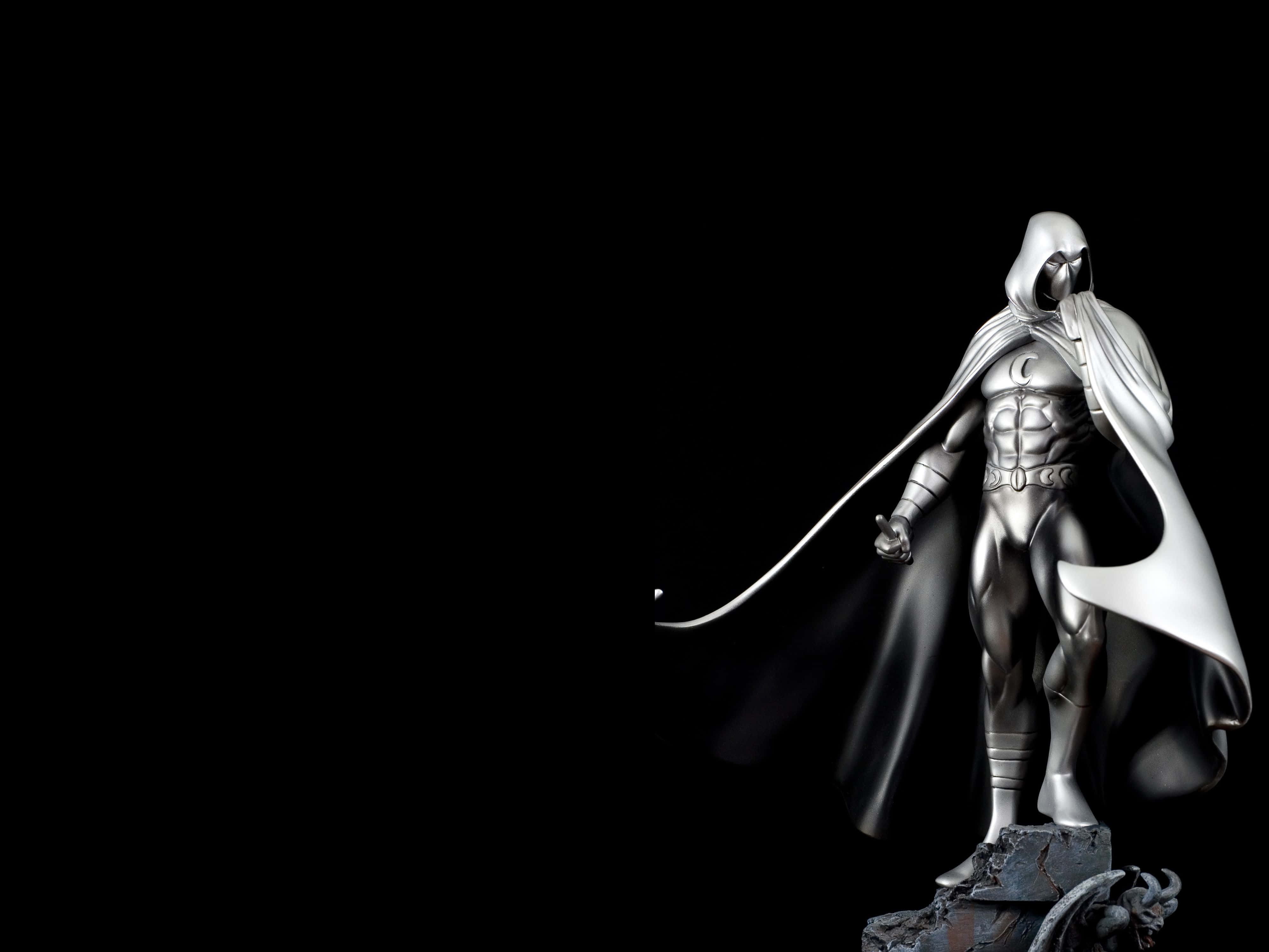 Moon Knight 桌布 壁紙 — 一個光滑的銀色兜帽超級英雄雕像，披著飄逸的斗篷，站在岩石底座上，背景是純黑色。