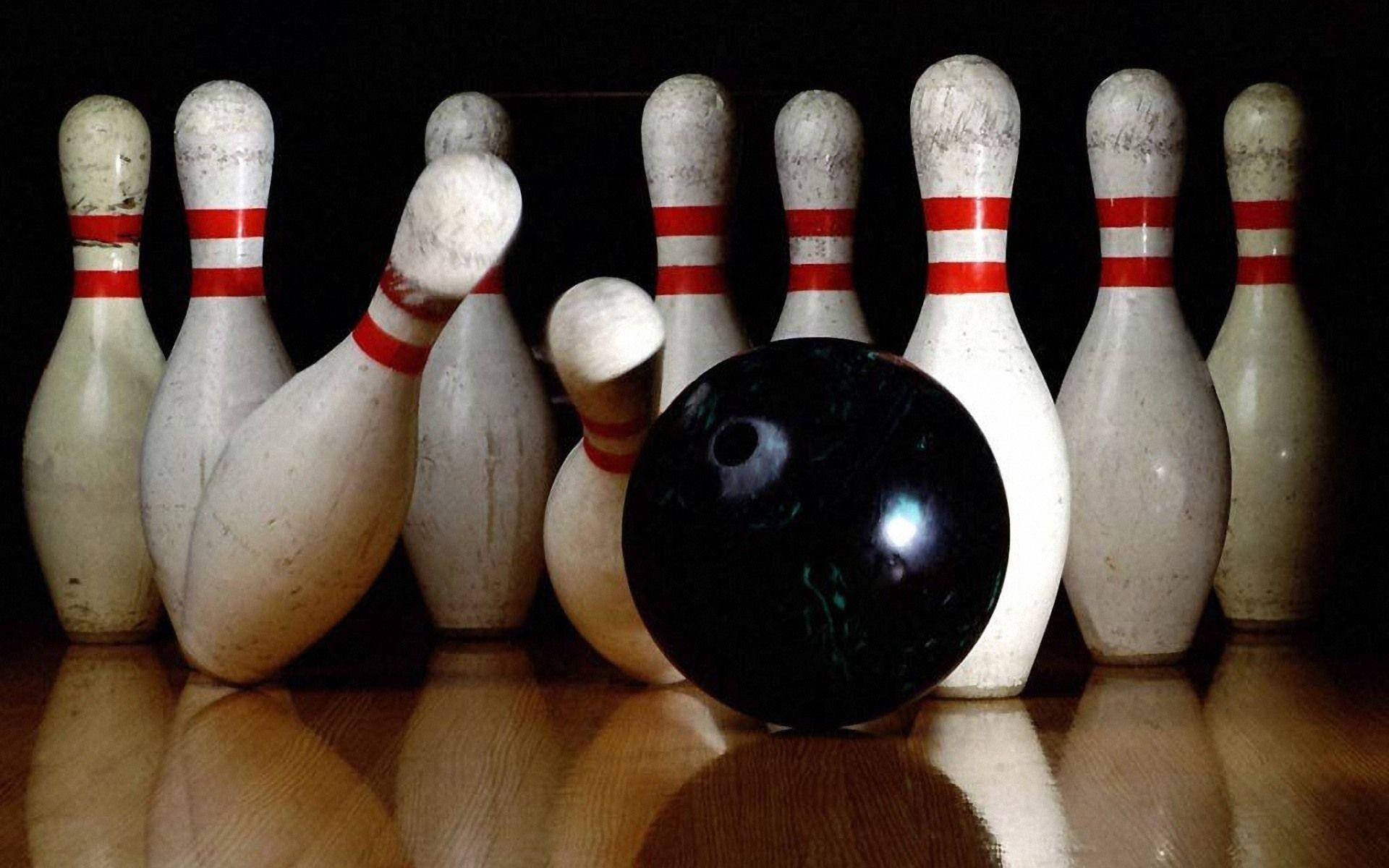 bowling pin 桌布 壁紙 — 一個光滑的黑色保齡球放在拋光木製球道上，前面是一組帶有紅色條紋的白色球瓶。