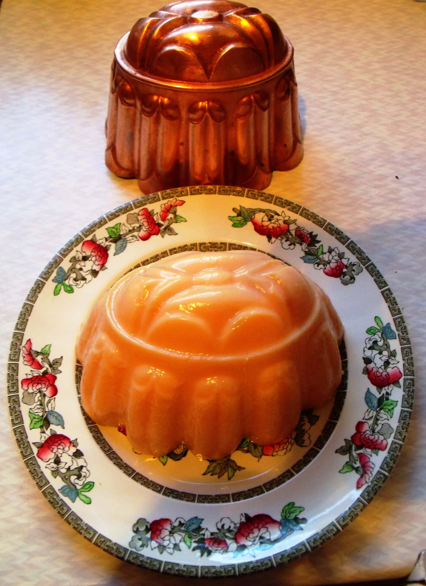 Blancmange 桌布 壁紙 — 一個光澤的桃色果凍模具放在一個花卉圖案的盤子上，旁邊是其銅製模具，在溫暖的室內燈光下。
