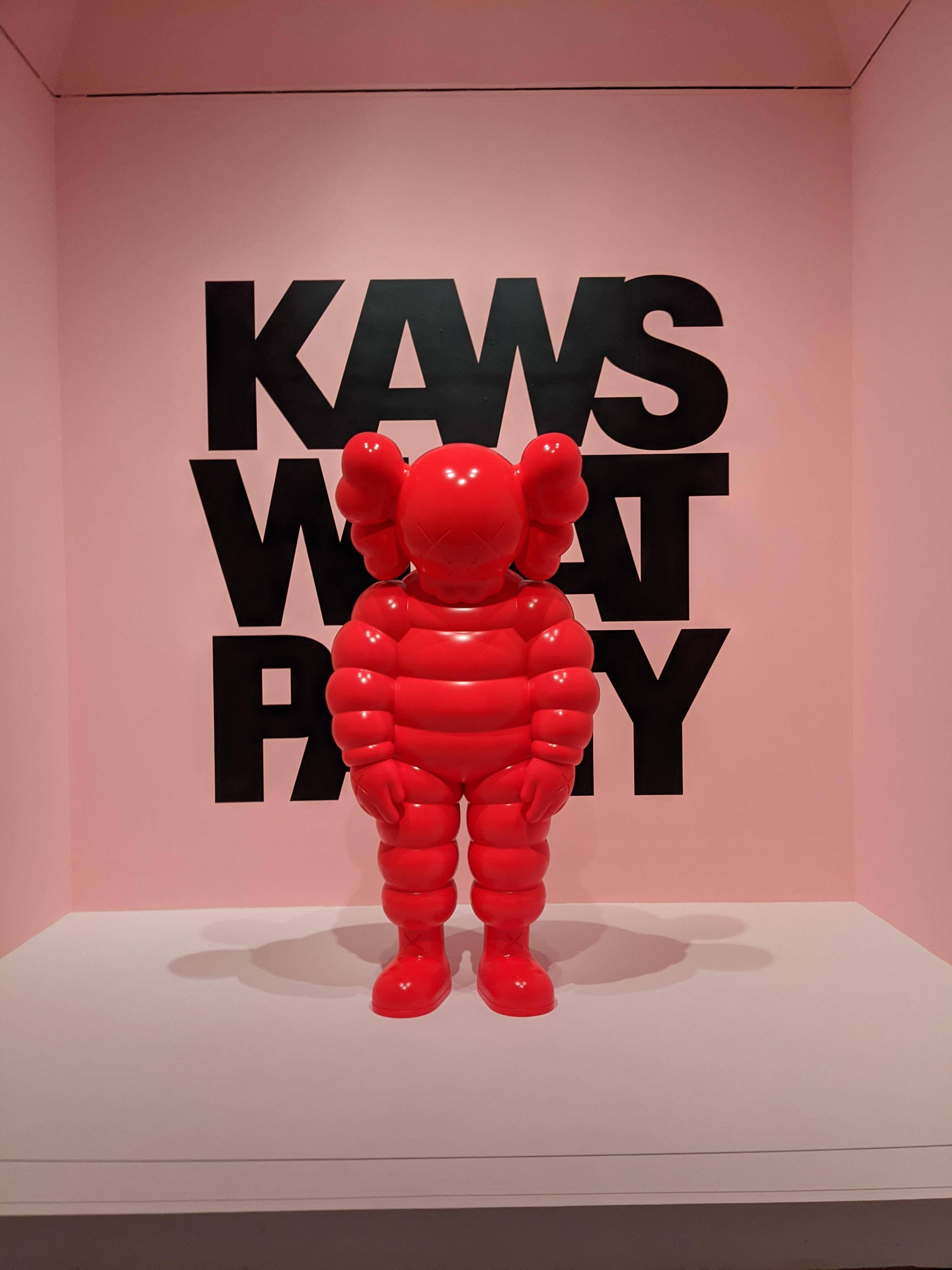 KAWS 桌布 壁紙 — 一個光澤的紅色KAWS Companion雕塑立在白色底座上，背景是粉紅色的牆壁，上面有粗體黑色'KAWS WHAT PARTY'文字。