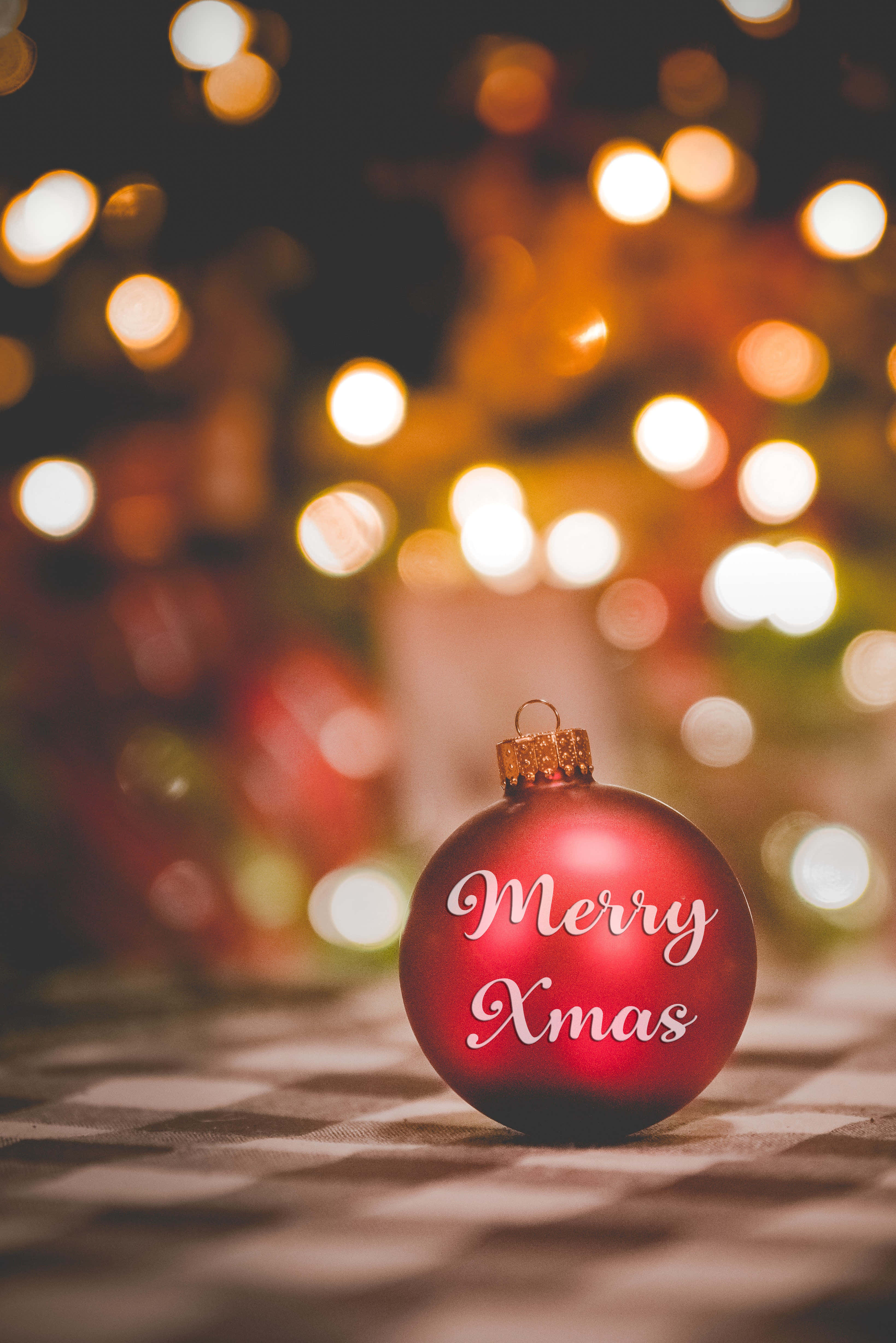 xmas 桌布 壁紙 — 一個光澤的紅色聖誕裝飾，上面有白色草書文字寫著Merry Xmas，放在格子表面上，背景是溫暖的散景節日燈光。