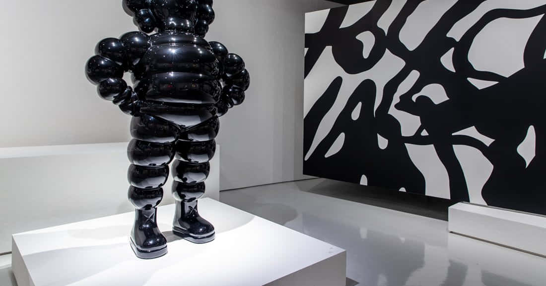 KAWS 桌布 壁紙 — 一個光澤的黑色氣泡形雕塑放在白色底座上，位於極簡主義畫廊中，旁邊是一幅大膽的黑白抽象牆面作品。