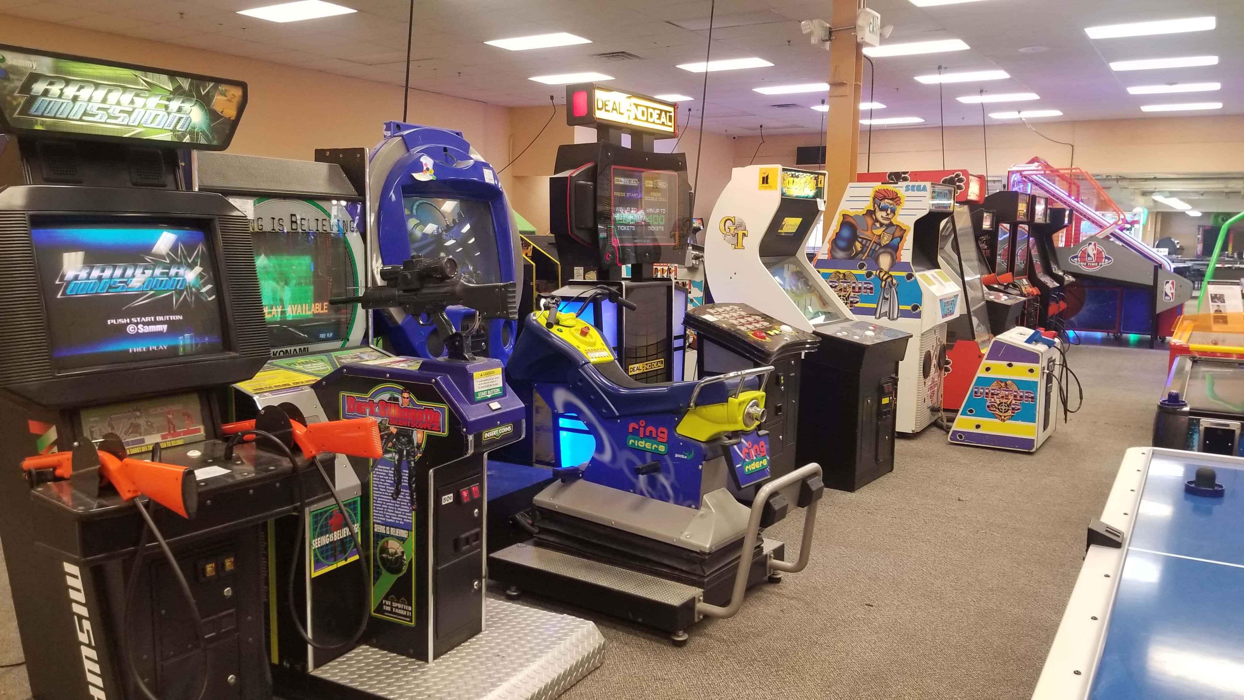 Arcade Games 桌布 壁紙 — 一個光線明亮的遊戲機中心，充滿了色彩繽紛的復古遊戲機和賽車模擬器，在螢光燈下閃耀。