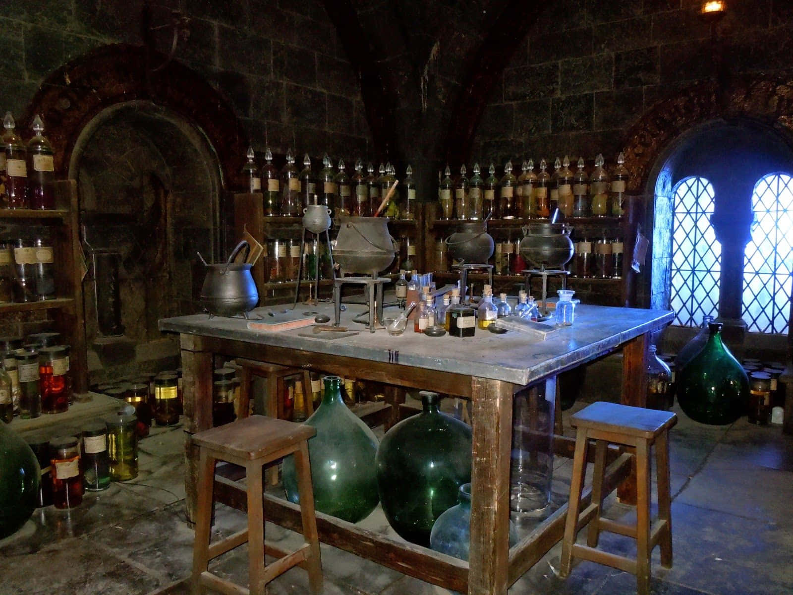 The Hogwarts Potions Class 桌布 壁紙 — 一個光線昏暗的中世紀石造實驗室，充滿冒泡的大鍋和藥水瓶架，在彩色玻璃光下。