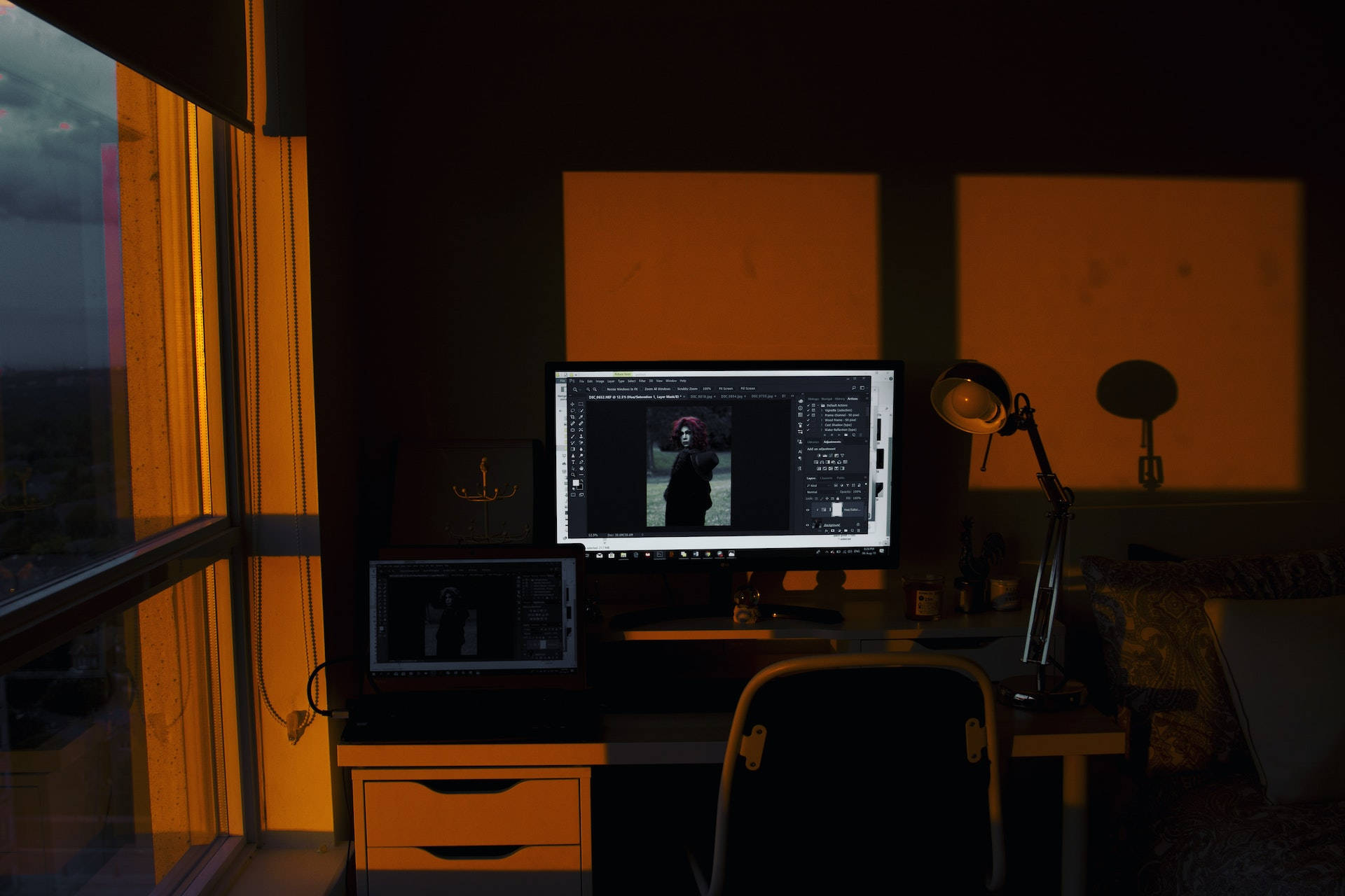 Office Space 桌布 壁紙 — 一個光線昏暗的家庭辦公室，沐浴在窗戶和檯燈散發的溫暖琥珀色光線中，電腦螢幕顯示著照片編輯軟體。
