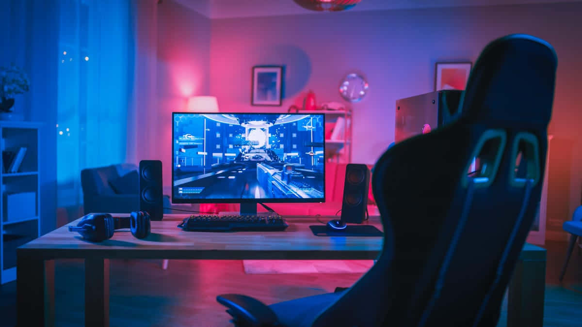 Gaming Pc Setup 桌布 壁紙 — 一個光線昏暗的遊戲室，沐浴在藍色和粉紅色的霓虹光中，顯示器上顯示賽車遊戲，前景有一張黑色人體工學椅。
