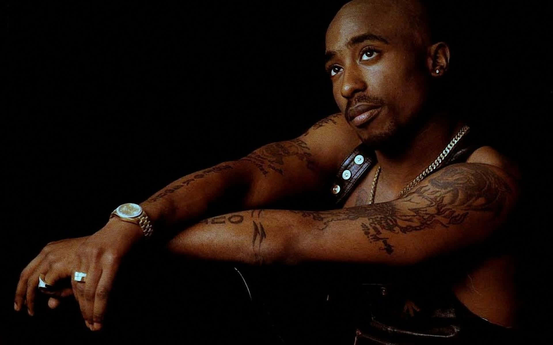 tupac 桌布 壁紙 — 一個光頭、手臂有刺青的男人，在黑色背景前用手托住下巴，戴著手錶和鏈條。