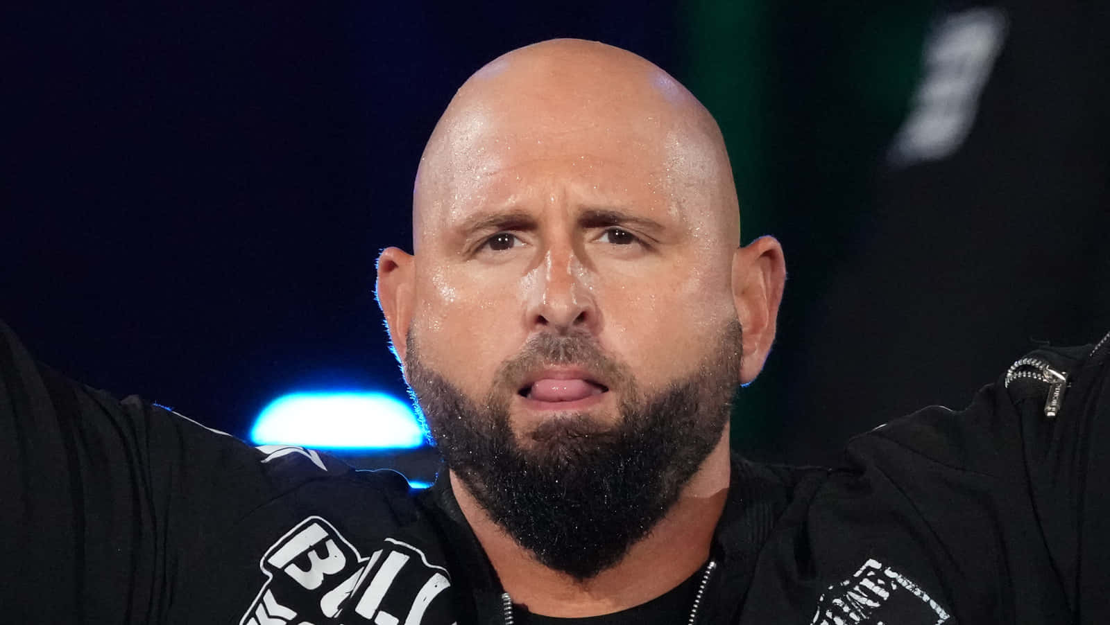 WWE 桌布 壁紙 — 一個光頭、濃密鬍鬚、表情強烈的男人，在明亮的舞台燈光下穿著一件帶有白色字樣的黑色外套。