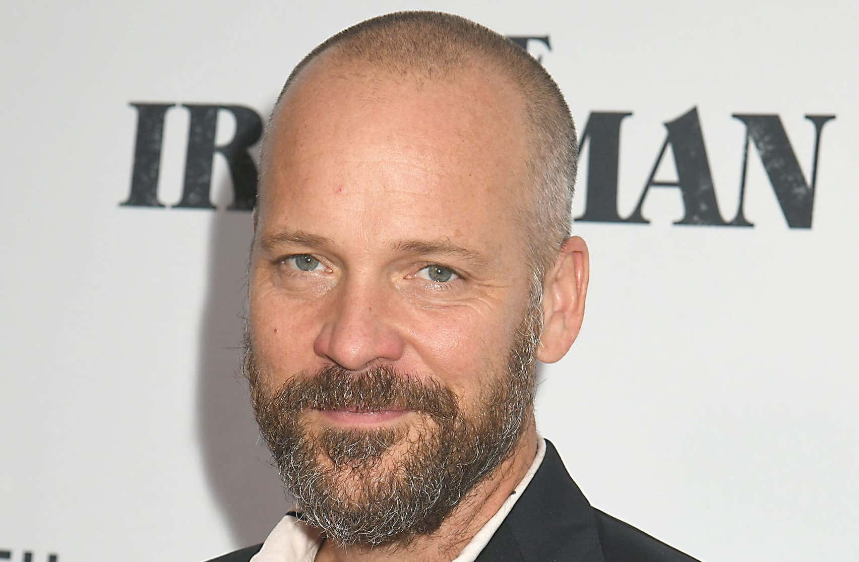 Peter Sarsgaard 桌布 壁紙 — 一個光頭、灰鬍鬚、身穿黑色西裝的男士特寫肖像，背景為白色，並有粗體文字。