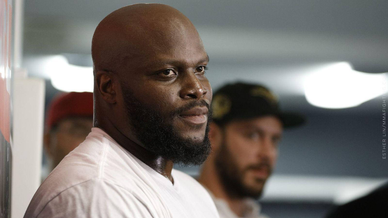 UFC 桌布 壁紙 — 一個光頭、留著滿臉鬍鬚、身穿白色T恤的男人，在柔和光線的室內環境中若有所思地望向一旁。