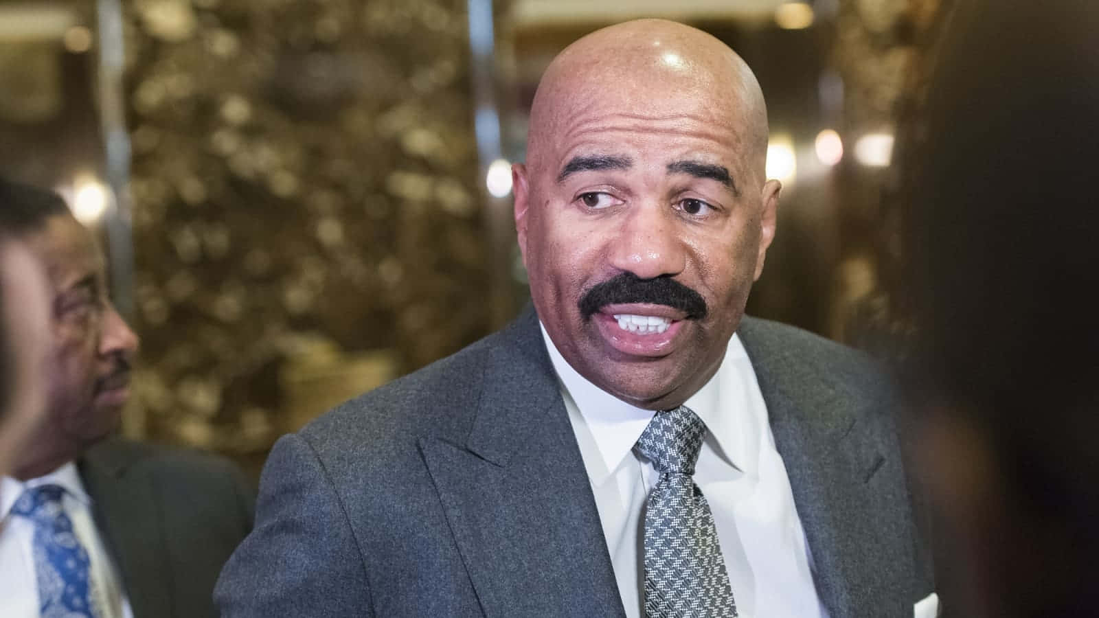steve harvey 桌布 壁紙 — 一個光頭、留著濃密鬍子的男人，穿著灰色西裝和圖案領帶，在金色紋理牆壁的室內微笑說話。
