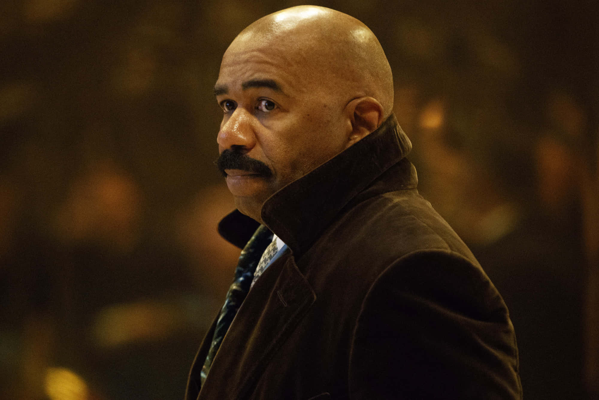 steve harvey 桌布 壁紙 — 一個光頭、留著濃密鬍子的男人，穿著深色外套，在溫暖照明的室內環境中專注地看向鏡頭外。