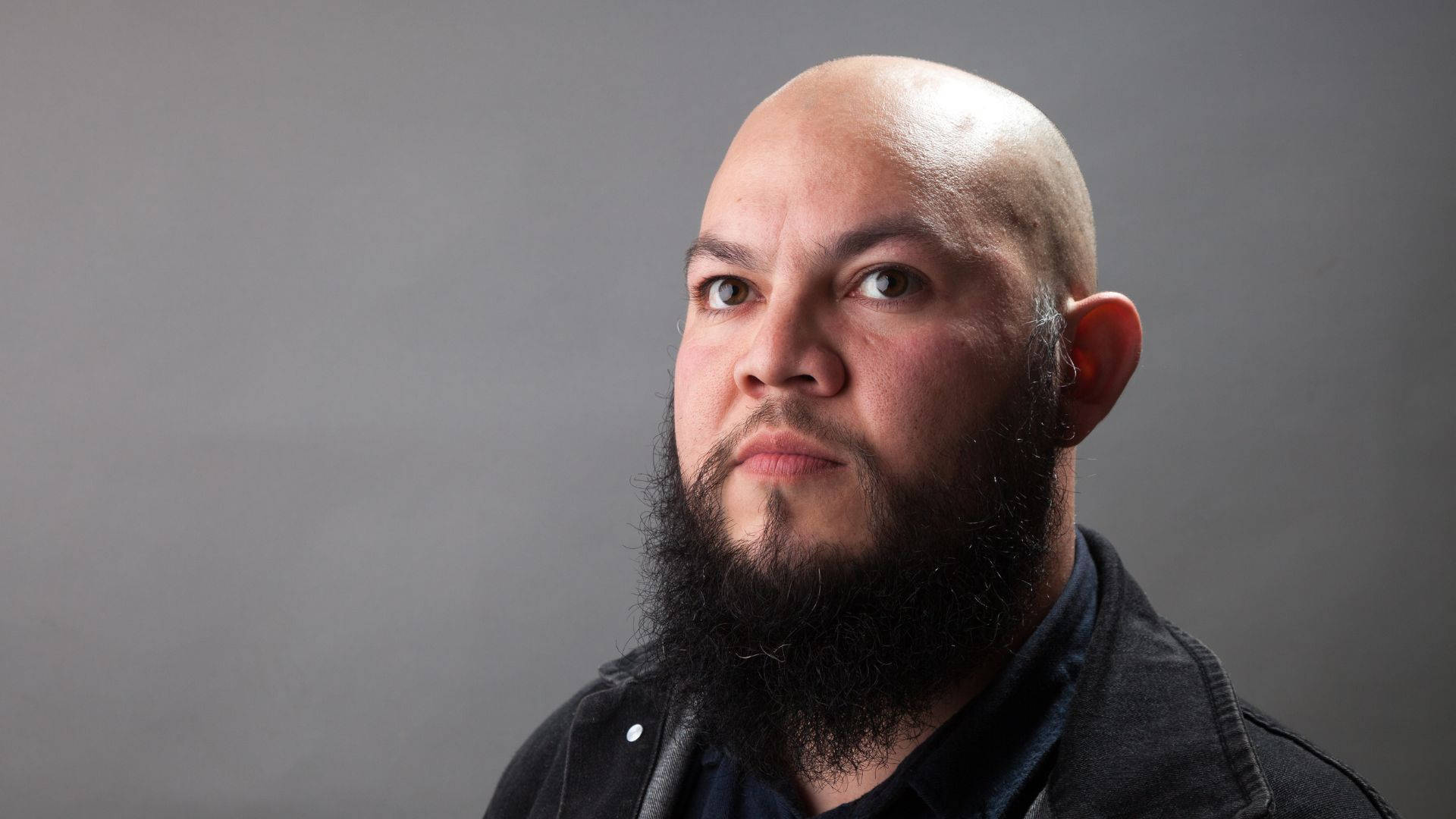 beard 桌布 壁紙 — 一個光頭、留著濃密鬍鬚的男人，在樸素的灰色工作室背景下專注地凝視。