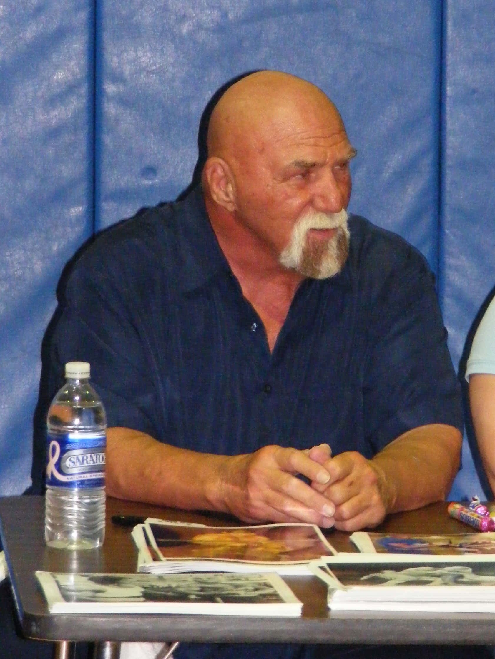 WWE 桌布 壁紙 — 一個光頭、留著白色山羊鬍的男人坐在桌前，在藍色背景前簽名。
