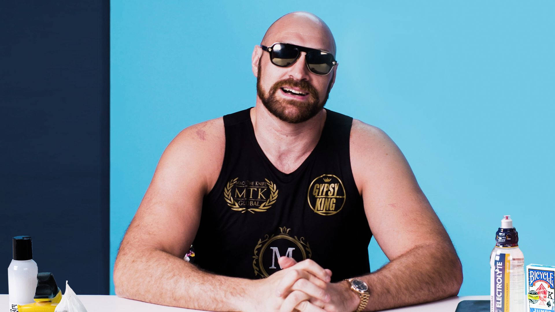 fury 桌布 壁紙 — 一個光頭、留鬍鬚、戴太陽眼鏡的男人坐在桌旁，身穿黑色背心，上面有金色標誌，背景是藍色和深藍色。