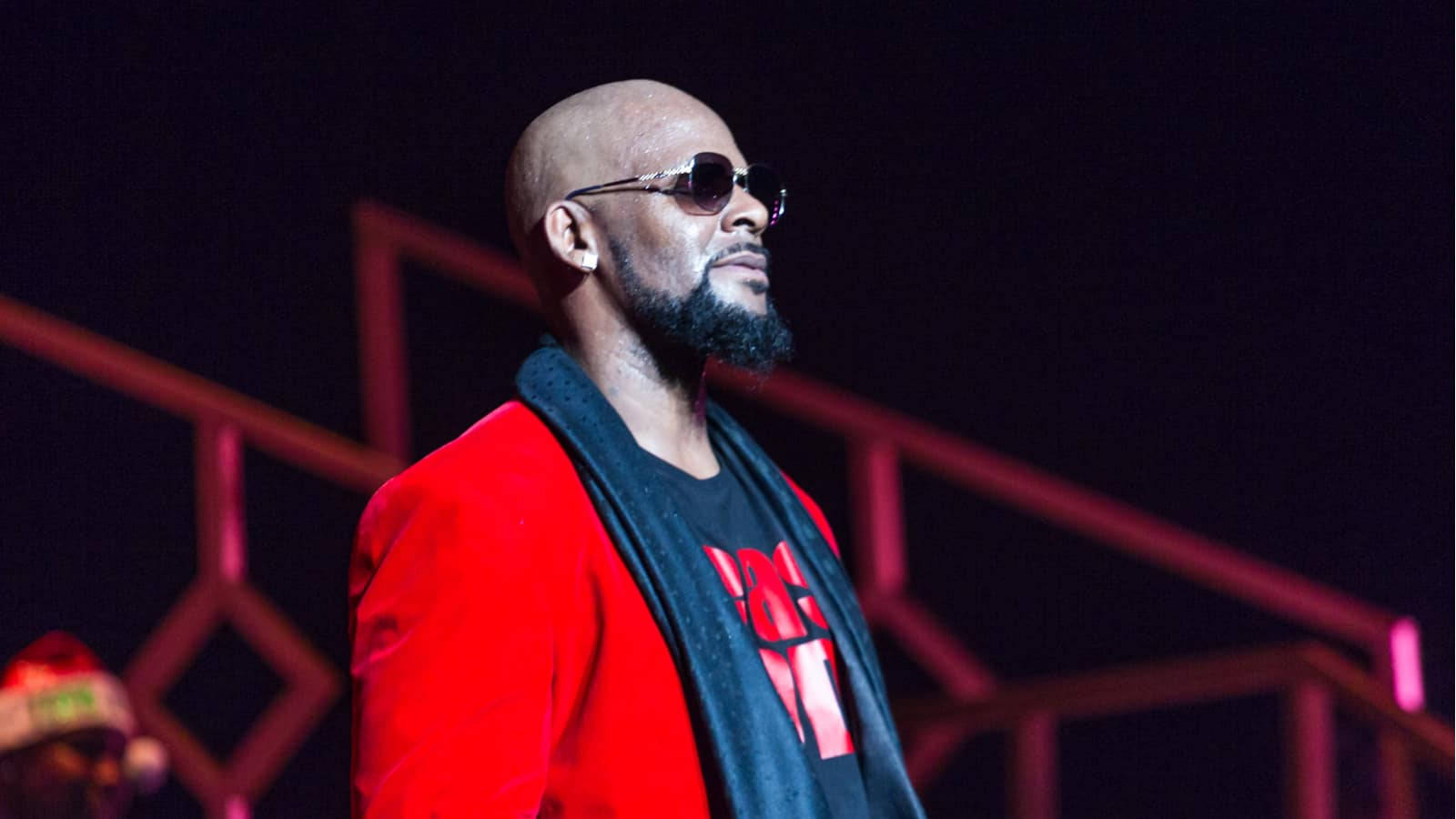 r kelly 桌布 壁紙 — 一個光頭、留鬍鬚、戴太陽眼鏡的男人，在舞台燈光下自信地穿著紅色外套。