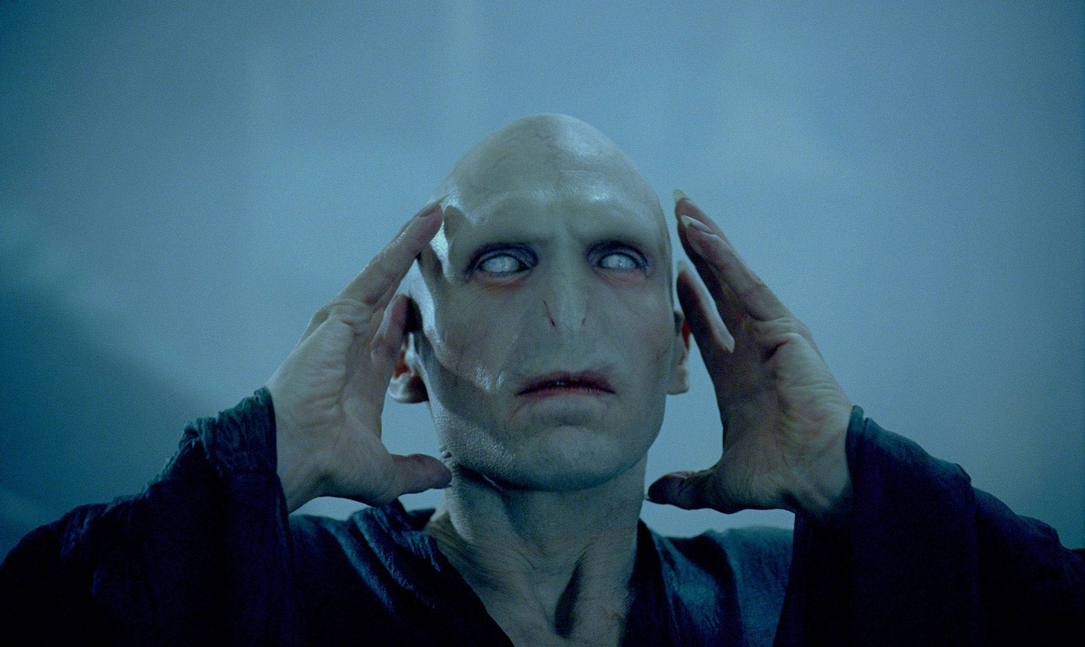 voldemort 桌布 壁紙 — 一個光頭、蒼白皮膚、大眼睛的男人，在模糊的藍色背景前，雙手按著太陽穴。