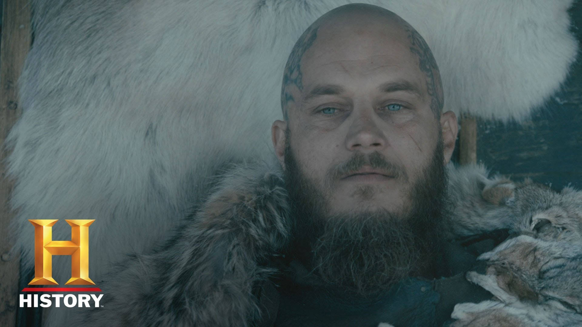 Vikings 桌布 壁紙 — 一個光頭、藍眼睛、臉上刺青的男人坐在毛皮襯裡的椅子上，專注地凝視著觀眾。