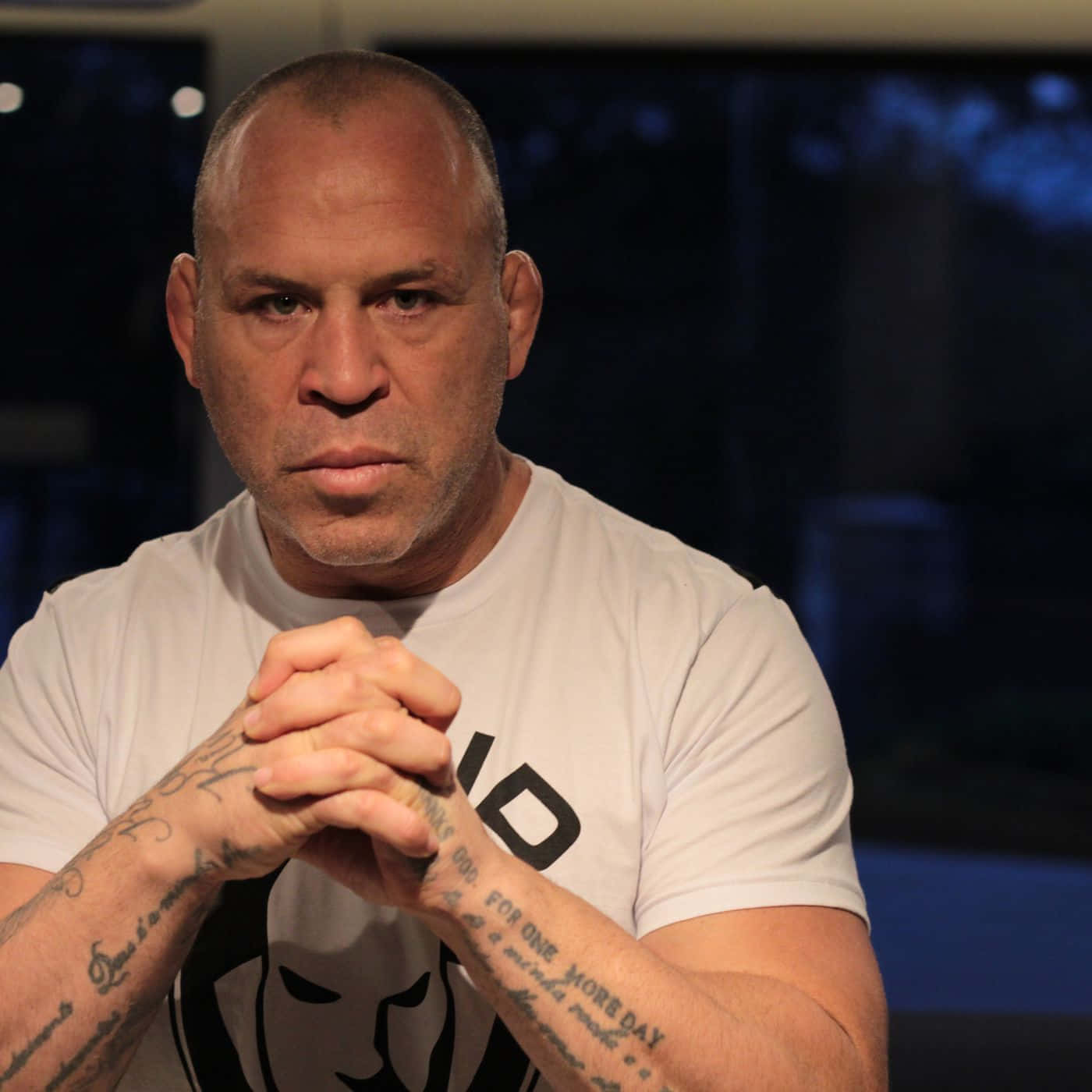 UFC 桌布 壁紙 — 一個光頭刺青男子雙手合十，身穿白色T恤，背景昏暗模糊。